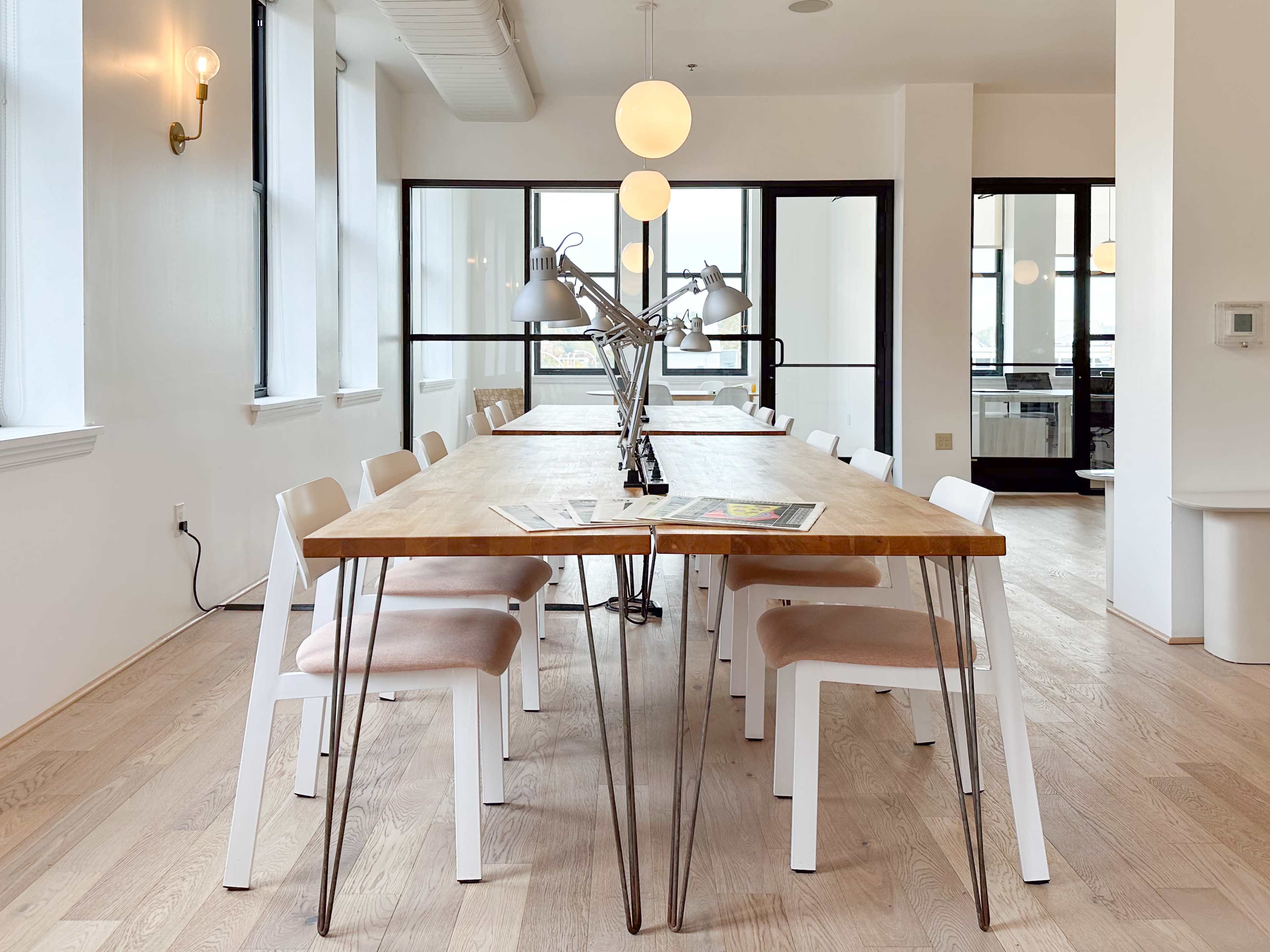 A long wooden table with metal legs and white chairs is set in a bright, minimalist room with large windows and modern lighting.