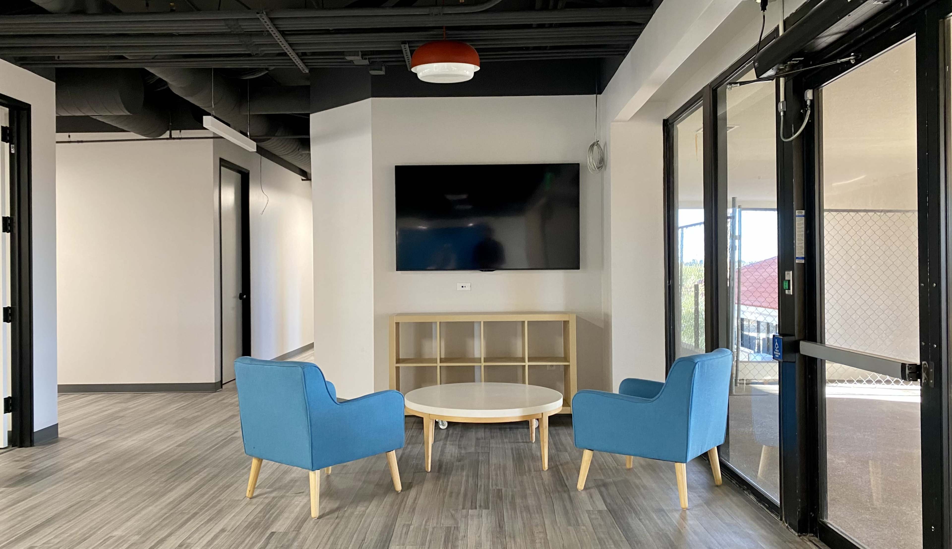 A small lounge area features two blue armchairs facing a coffee table in front of a wall-mounted television, with large windows allowing natural light to enter.