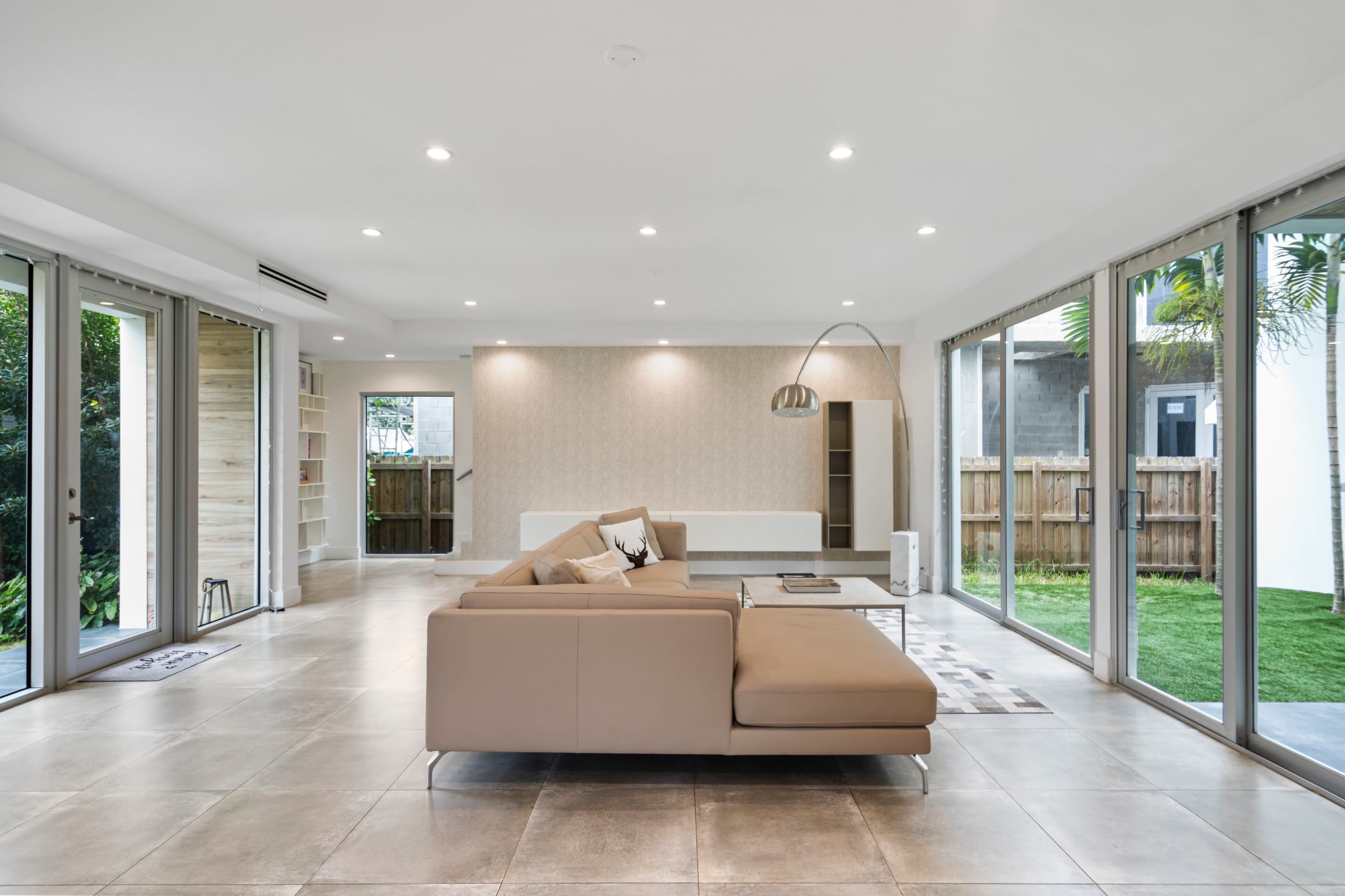 A modern living room with large glass doors, a beige sectional sofa, and a minimalist coffee table, surrounded by greenery.