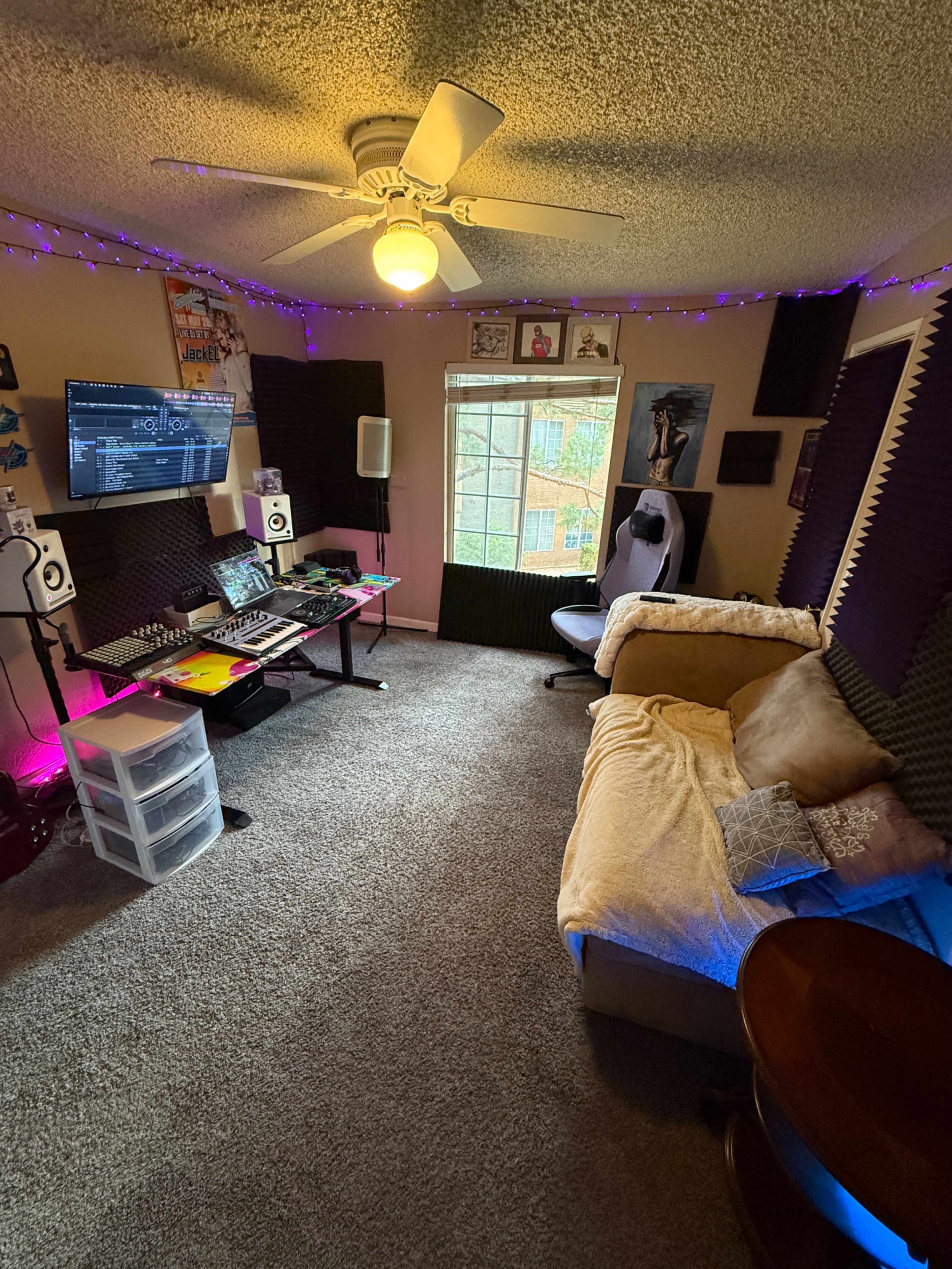 The image shows a small music studio or recording space, featuring a mixing console, computer monitor, and a cozy seating area with a couch and coffee table, all illuminated by string lights.