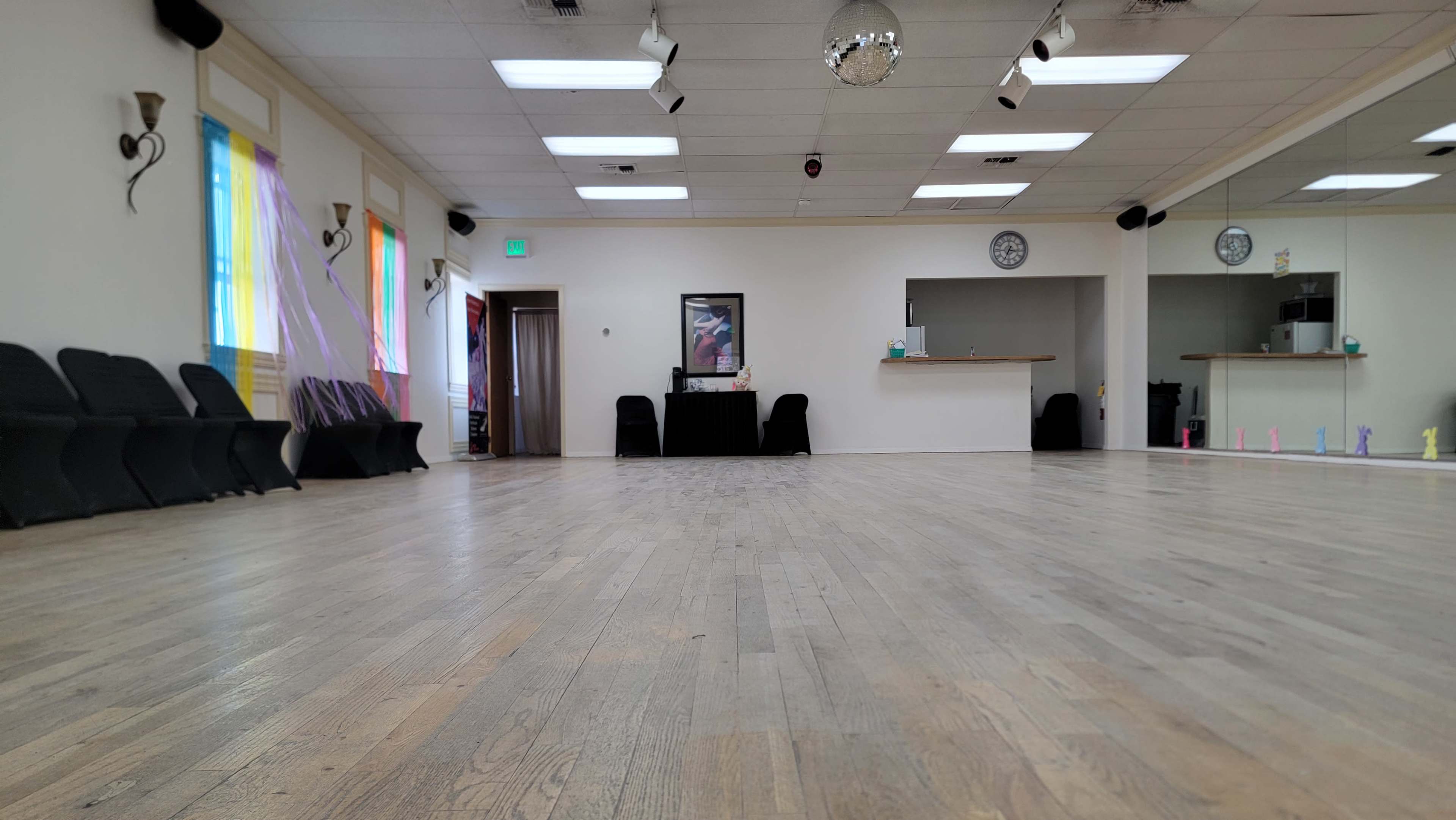 The image shows a spacious dance studio with wooden floors, illuminated by natural light from windows, featuring chairs along one wall and a small reception area in the background.