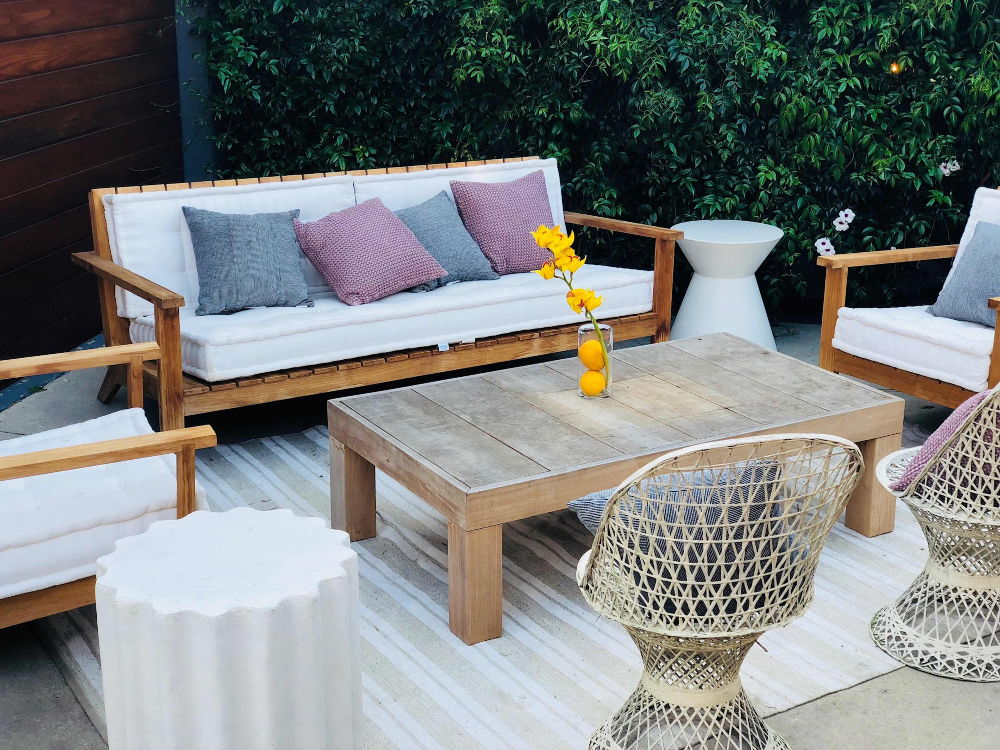 A patio seating area features wooden chairs and a table, with cushions and a flower vase, surrounded by greenery.
