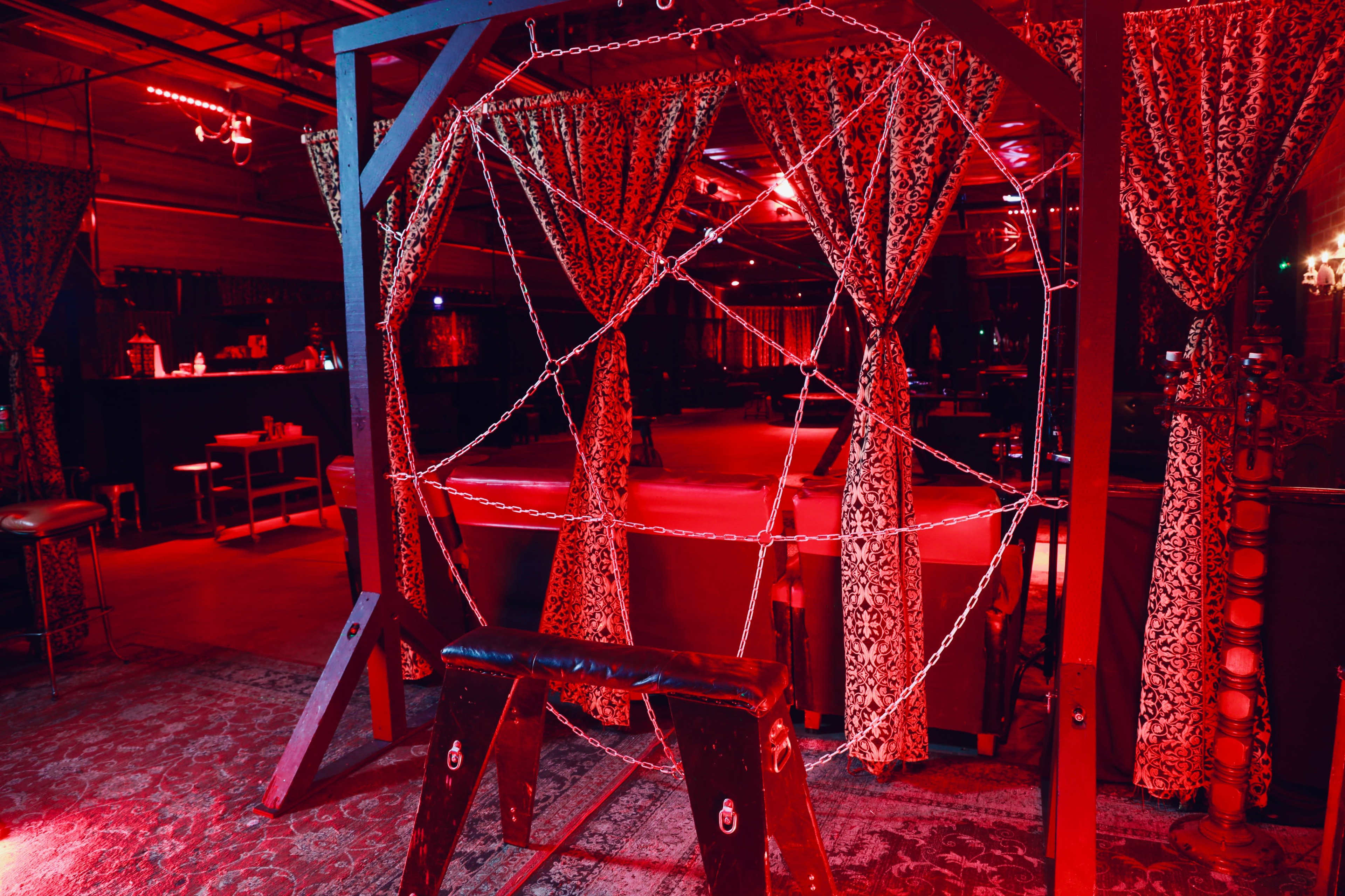 The image shows a dimly lit room with red lighting, featuring a large decorative frame adorned with curtains and chains in front of a seating area.