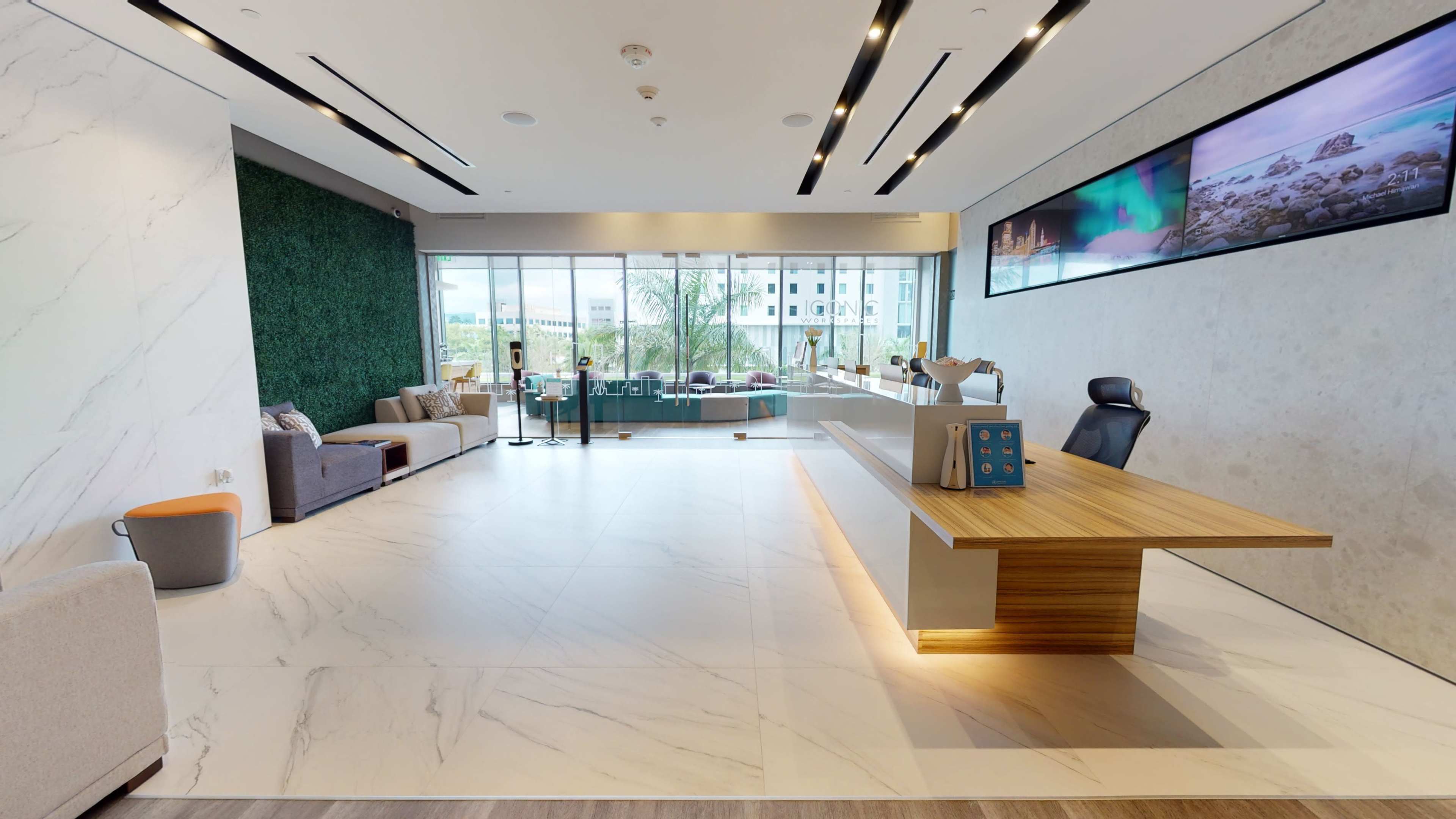 A modern office reception area with a large wooden front desk, a seating area with sofas, and large windows overlooking greenery.
