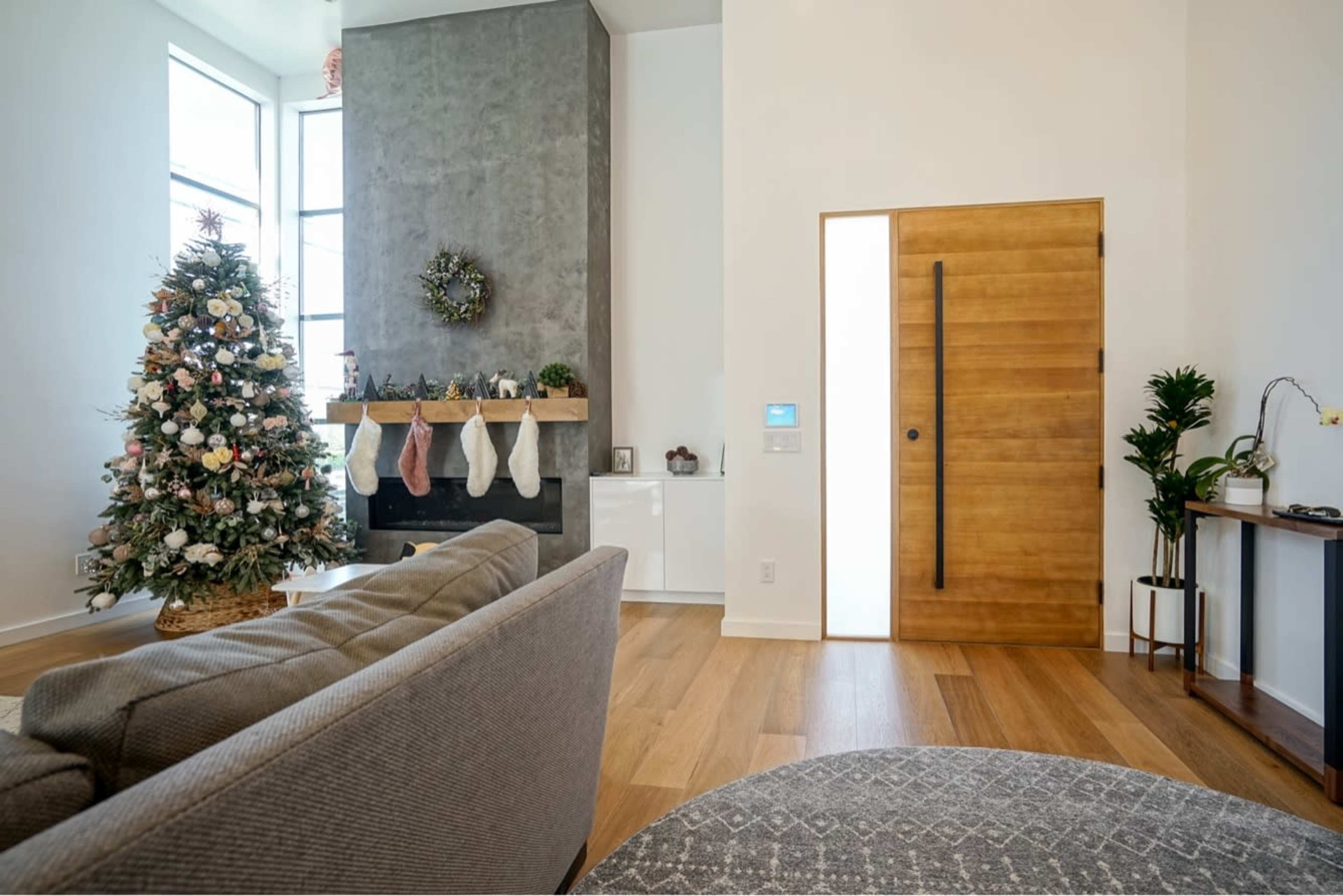 The image shows a modern living room featuring a decorated Christmas tree, a stone fireplace with stockings, and a wooden door leading to another space.