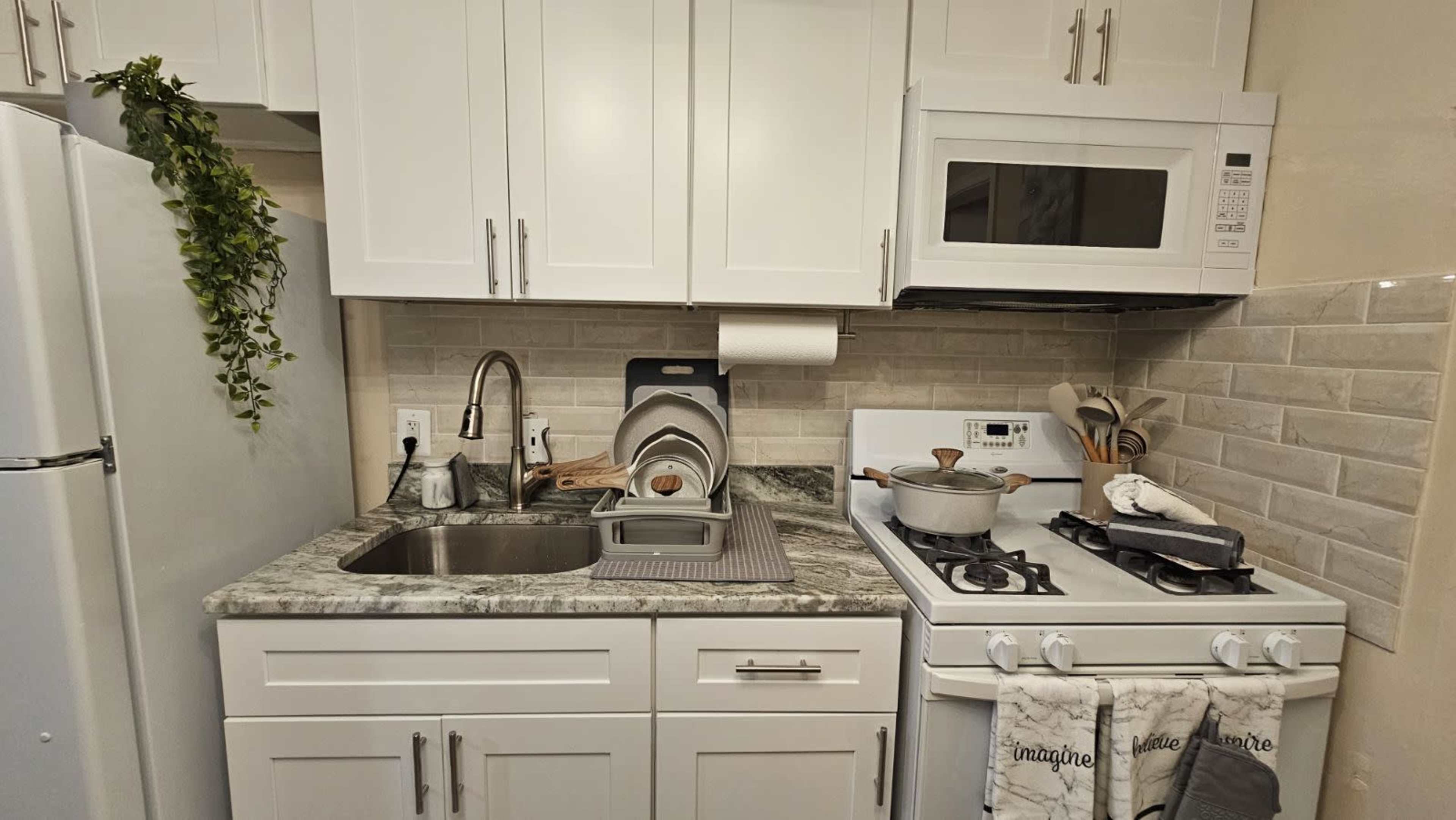 A modern kitchen with white cabinets, a sink, a stovetop, and a microwave, all featuring a stone countertop and gray tile backsplash.