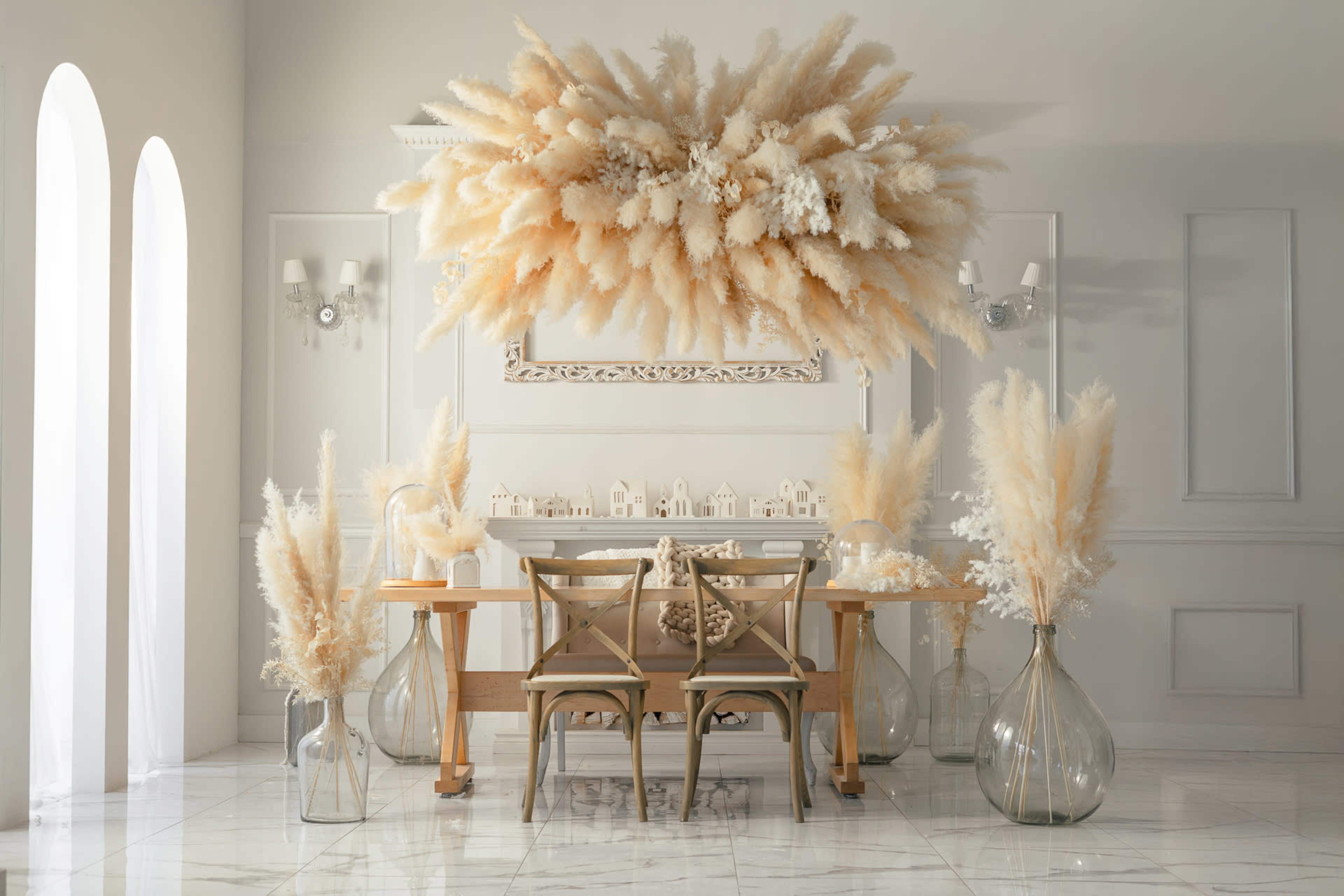 A minimalist dining room features a wooden table with two chairs, surrounded by decorative dried plants and a wall adorned with a large floral arrangement.