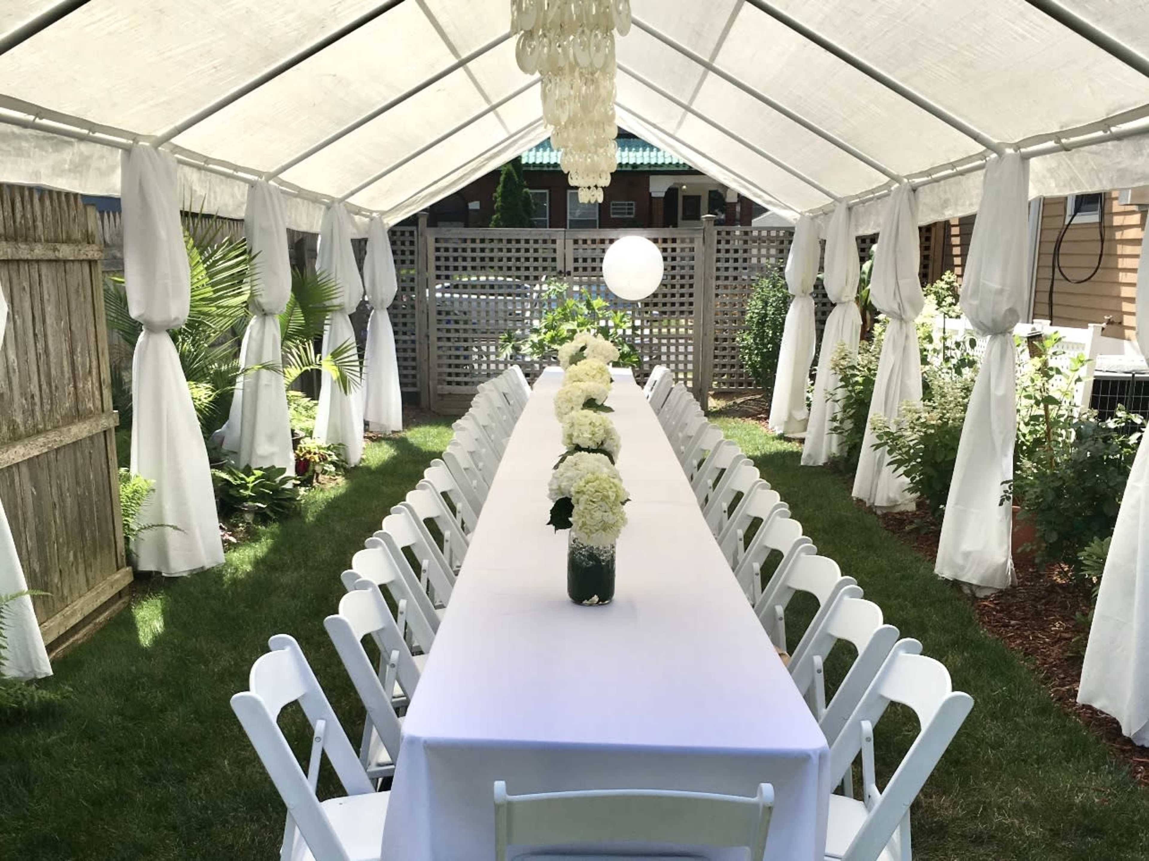 A long table is set under a white tent, adorned with floral centerpieces and surrounded by chairs on a grassy area.