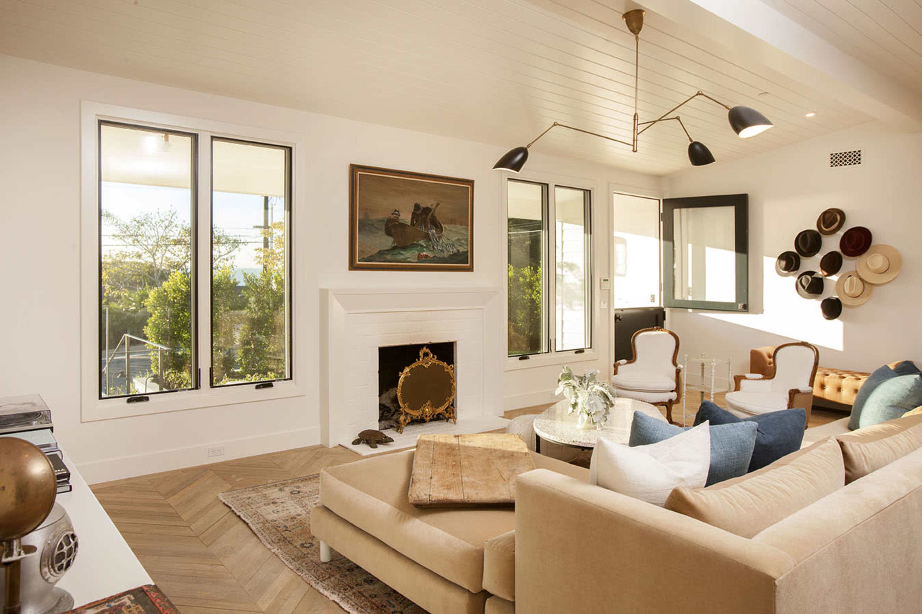 The image shows a modern living room with a beige sofa, a coffee table, and large windows framing a scenic view, complemented by a gallery wall of hats.