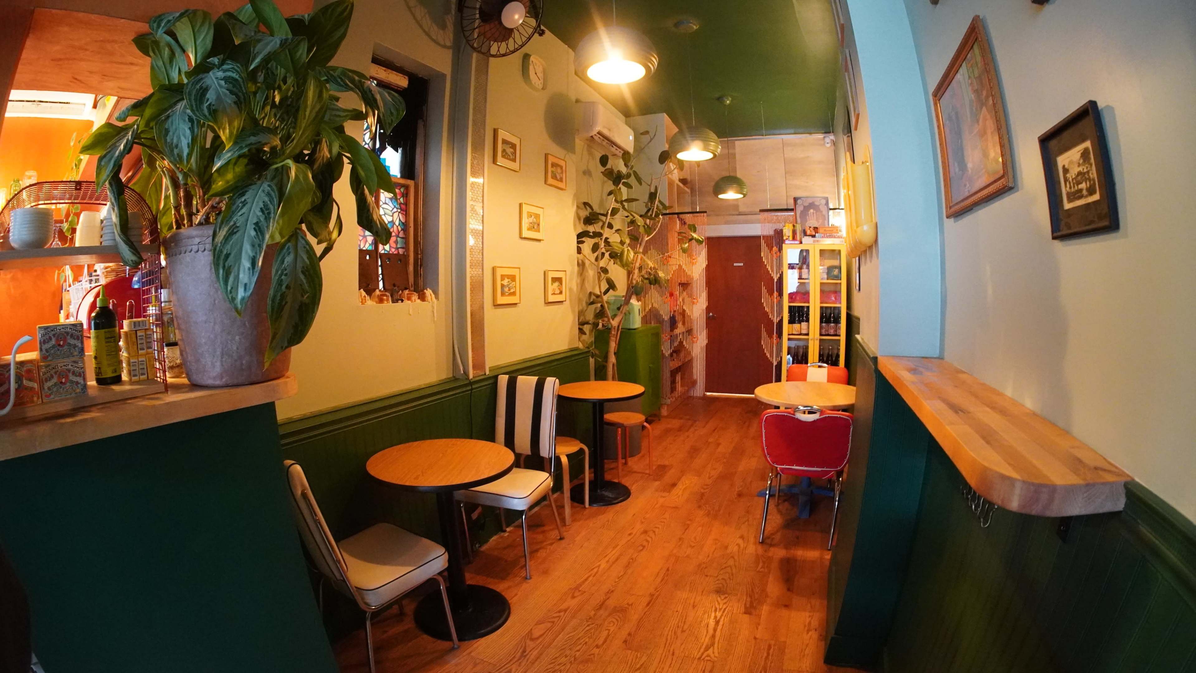 The image shows a narrow café interior with wooden flooring, green walls, small round tables, and a few chairs, along with plants and framed pictures on the walls.