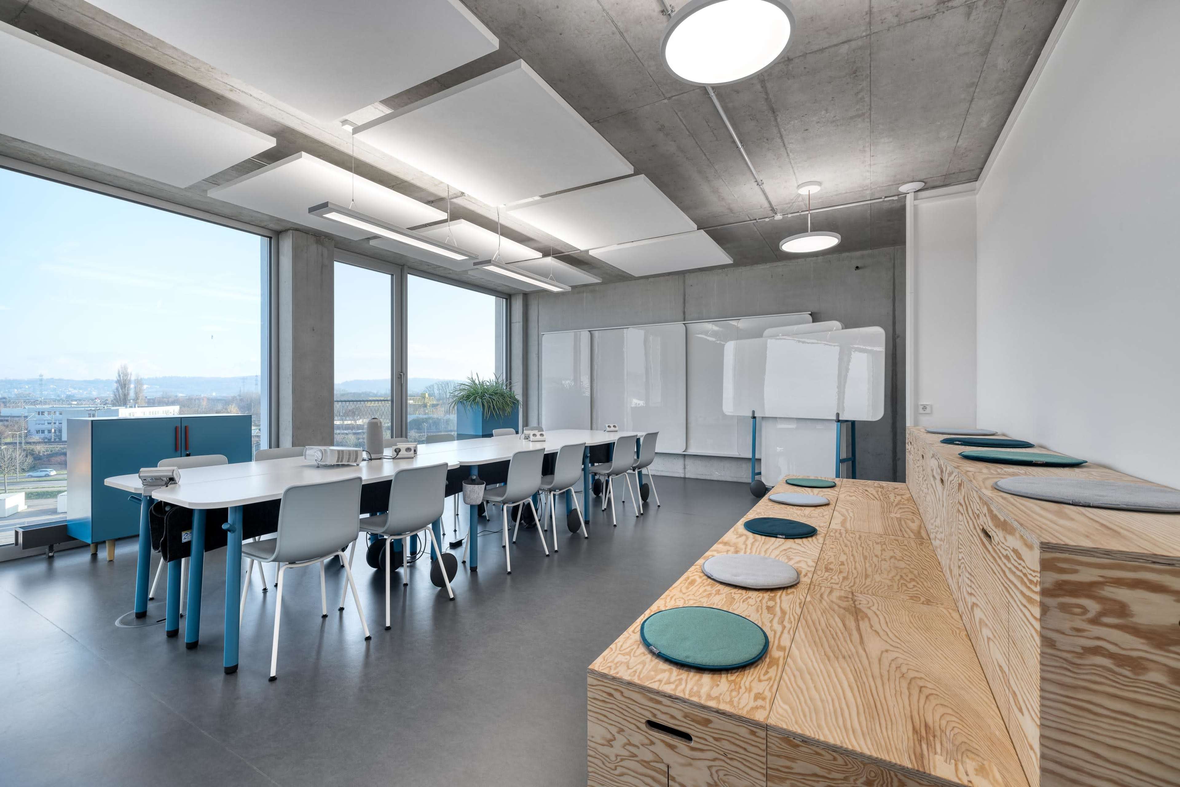 A modern office meeting room features a large table surrounded by chairs, with a wall of windows offering a view outside, and wooden benches along one side.