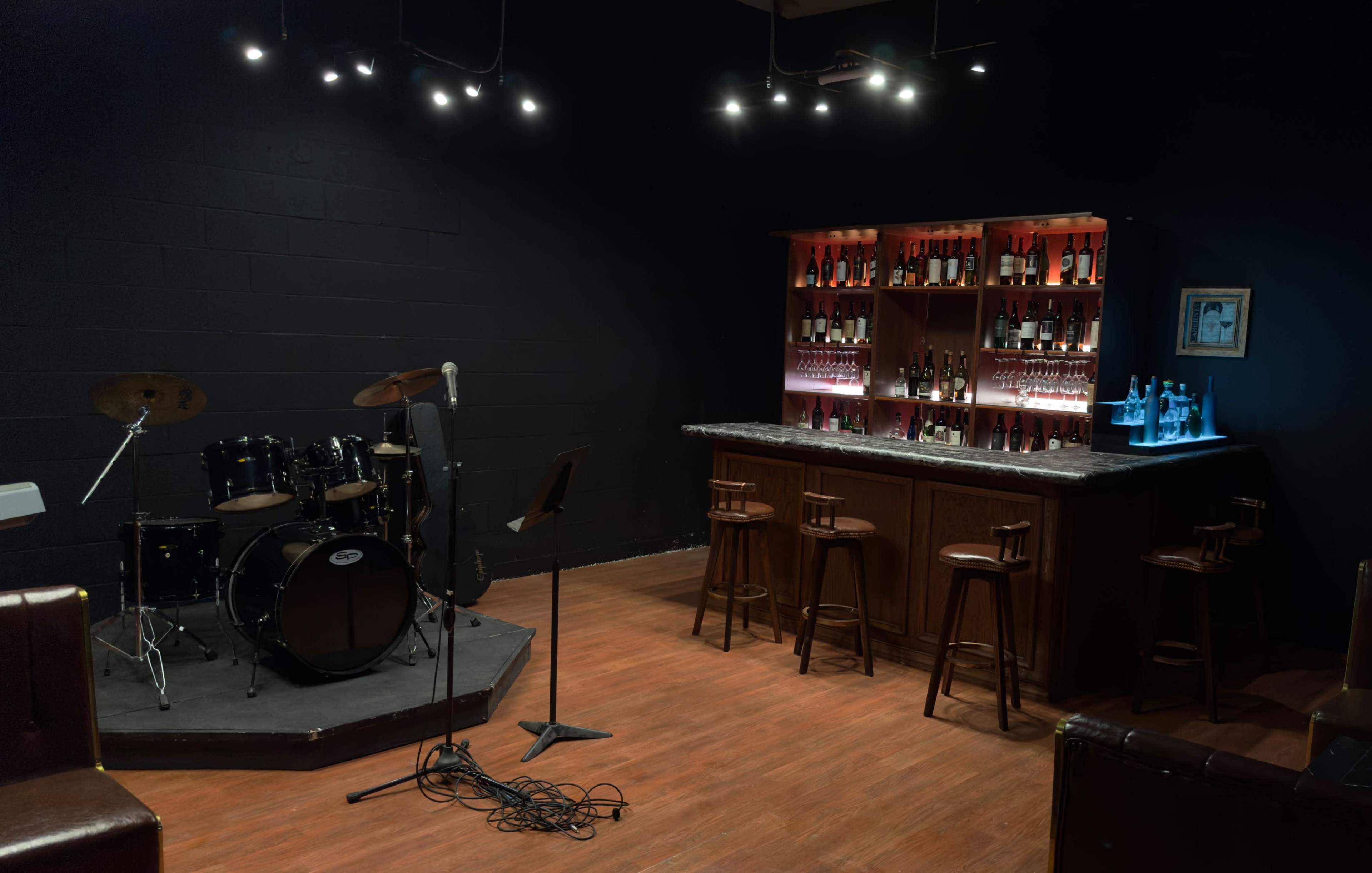 A small music venue features a drum set on a stage, a microphone stand, and a bar illuminated by shelves of bottles.