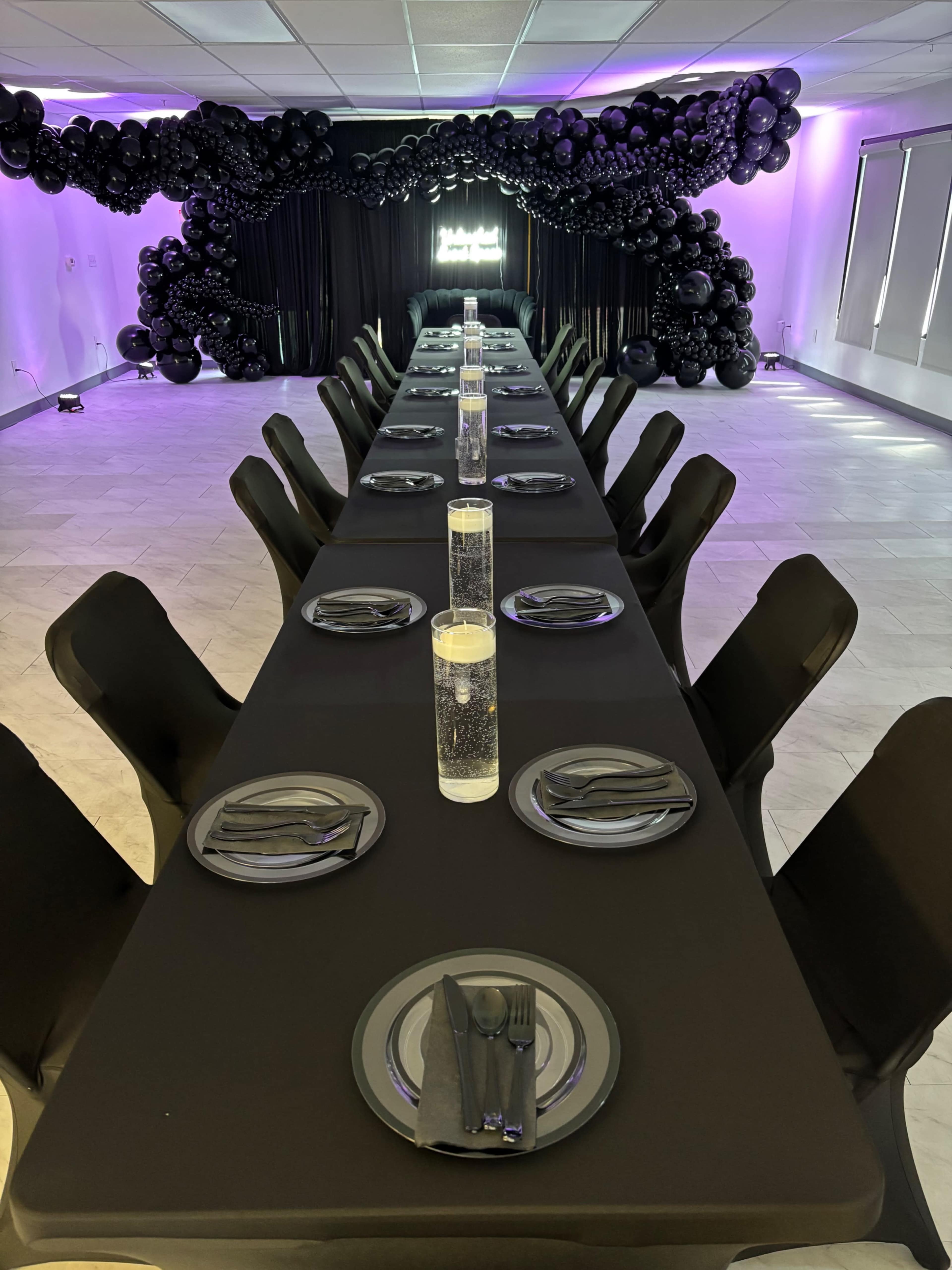 A long black table is set for a formal dinner, with clear glass vases filled with water and floating candles as centerpieces, and a backdrop of black balloons.
