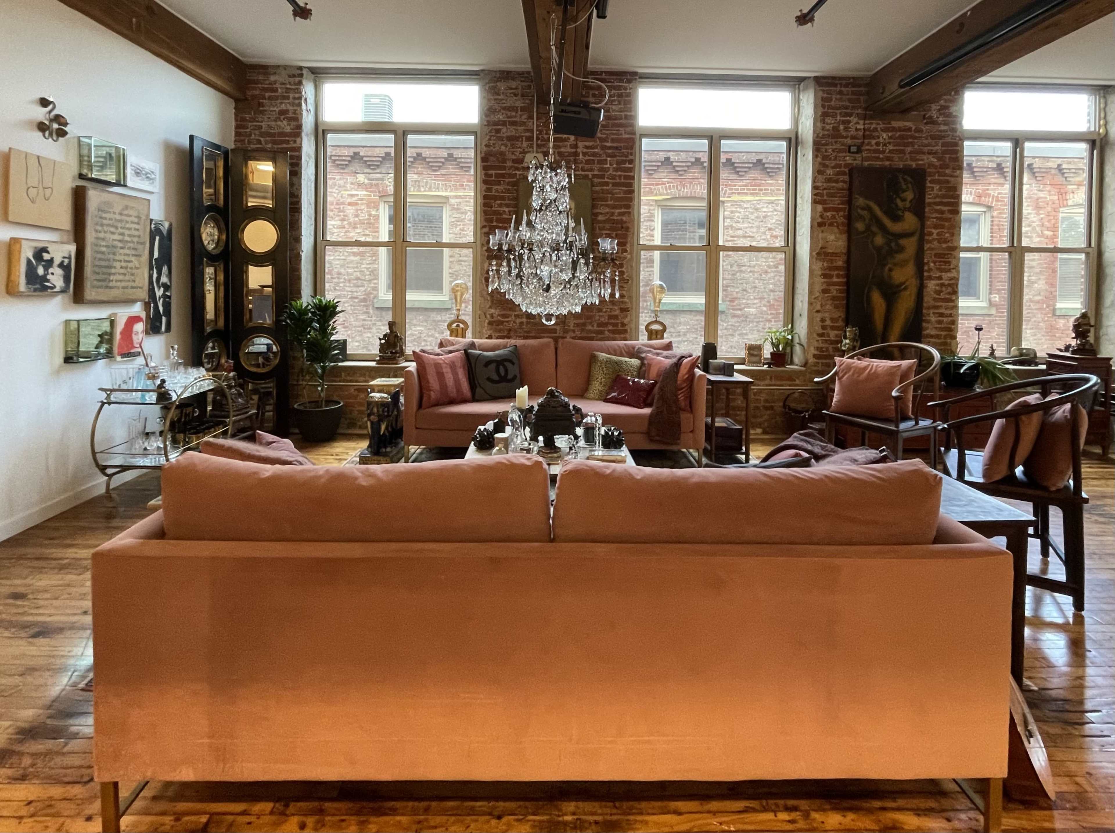 The image shows a stylish living room featuring a large, rose-colored sofa, a chandelier, and various pieces of artwork on exposed brick walls.