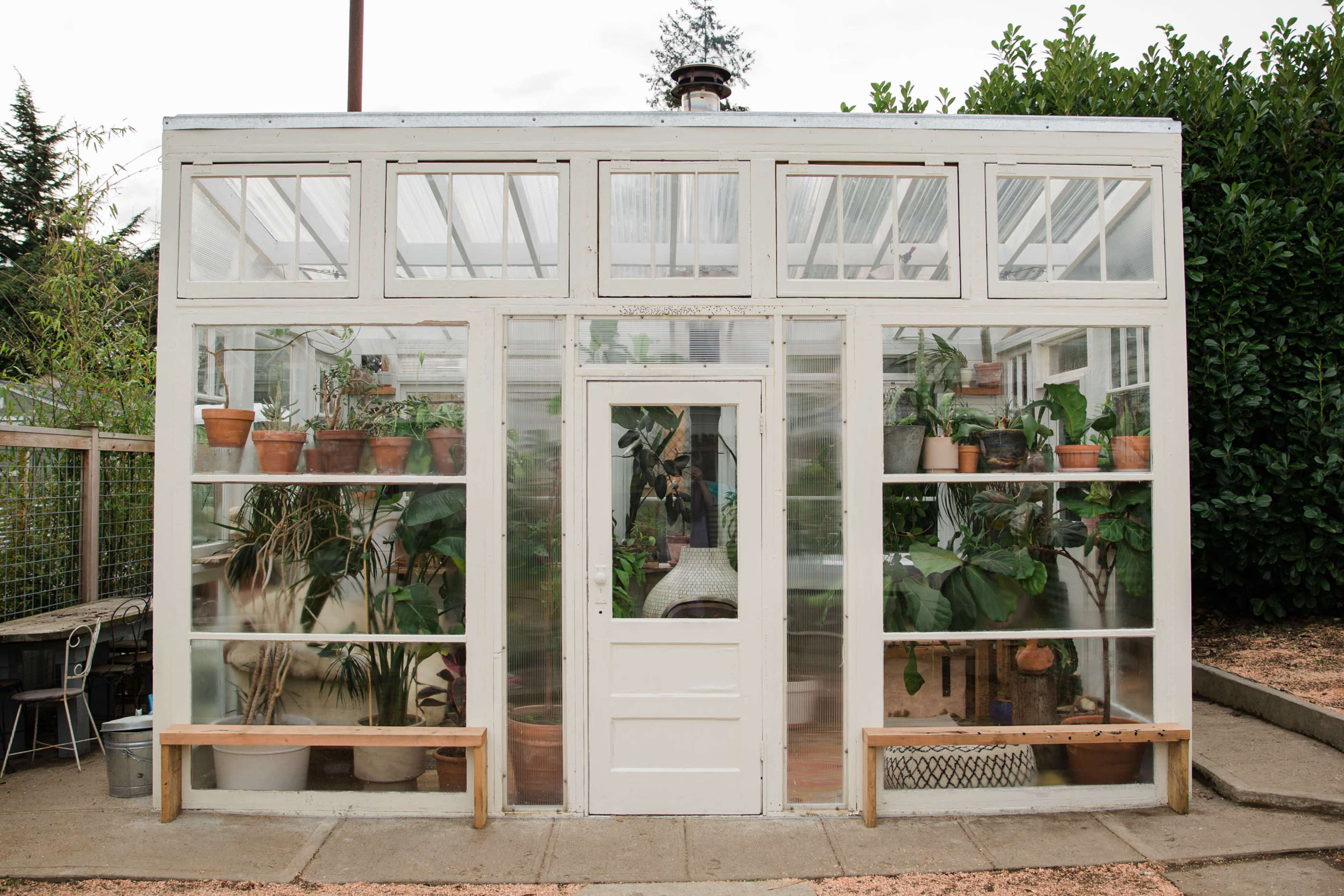 A white-framed greenhouse with glass panels, featuring potted plants on shelves and wooden benches outside.