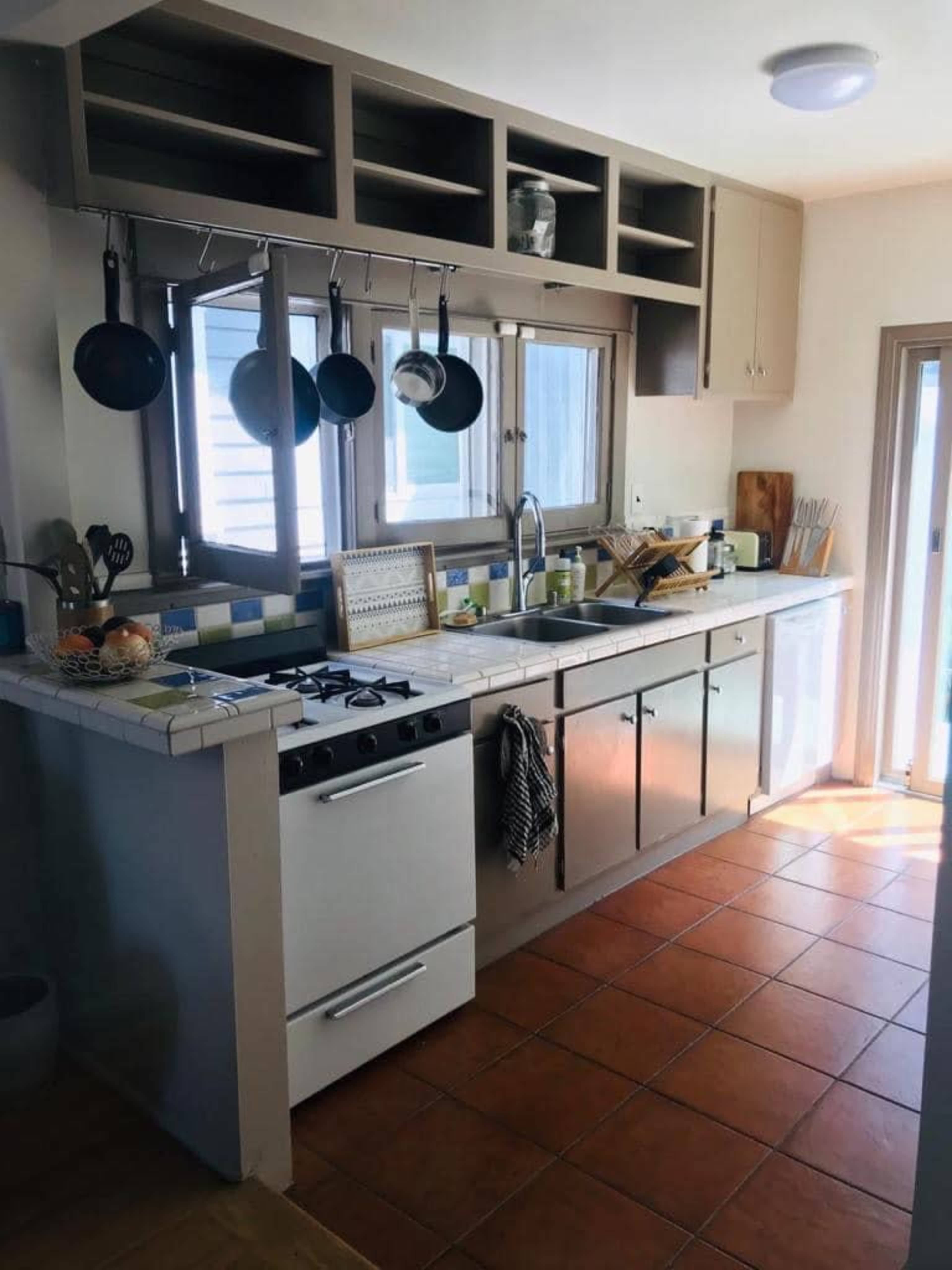 The image shows a bright kitchen featuring a tiled counter with a stove, hanging pots, and open shelves above, illuminated by natural light coming through a window.