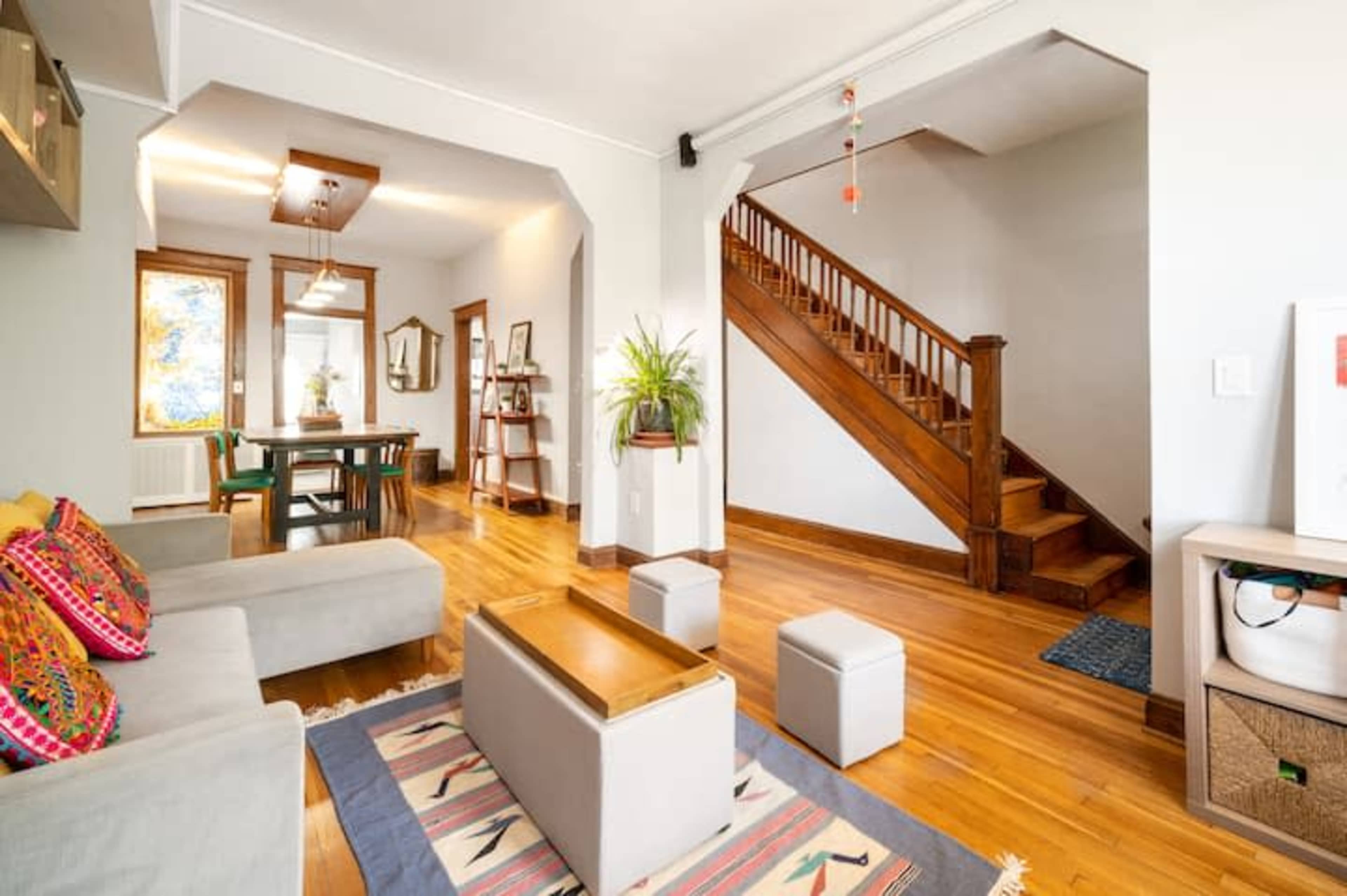A living room with a light-colored sofa, a wooden coffee table, and a staircase leading to an upper level, with a dining area visible in the background.