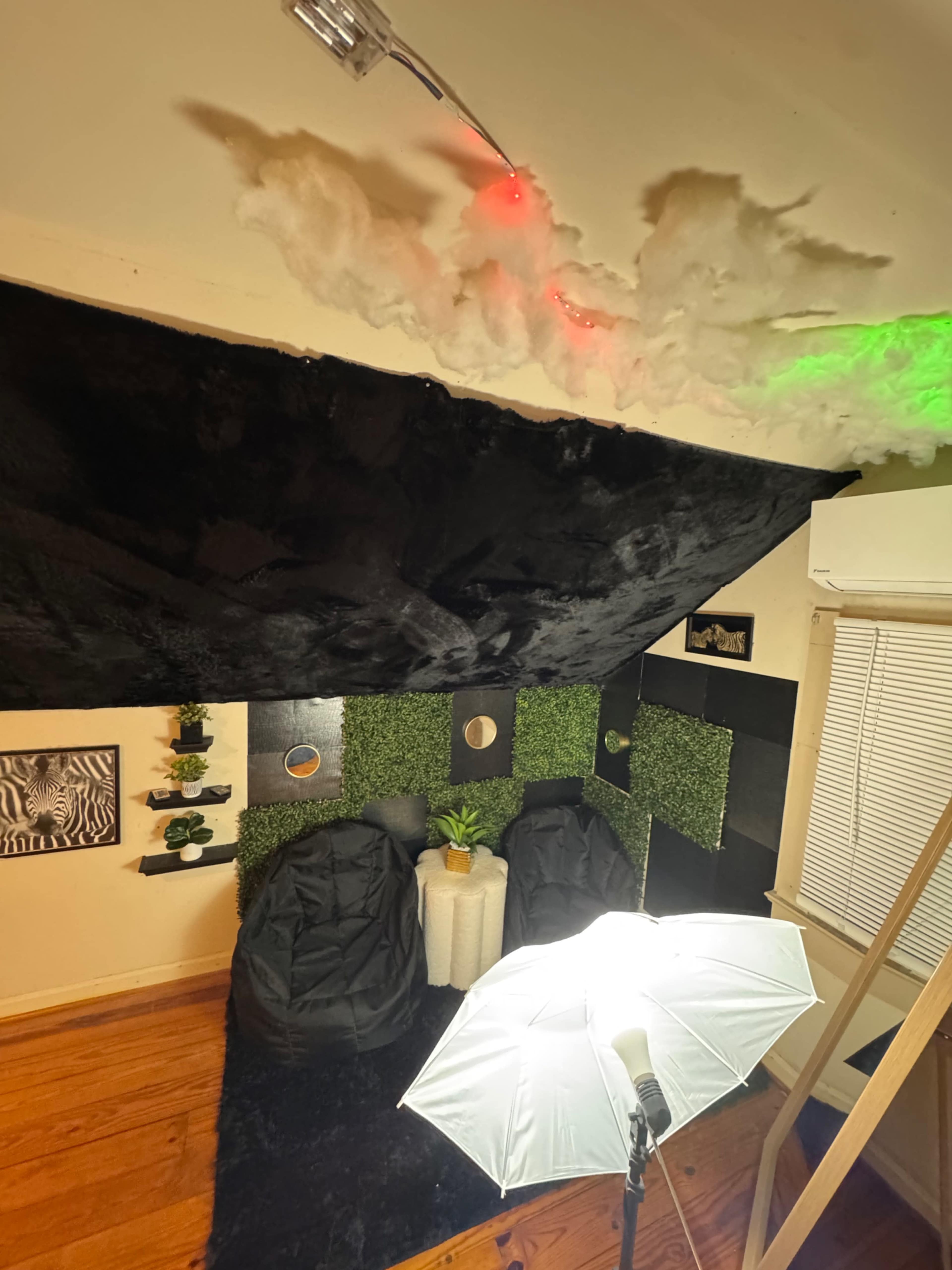 The image shows a creatively decorated room featuring a black wall with cloud-like decor, two bean bag chairs, and a small plant on a table, illuminated by a soft light source.