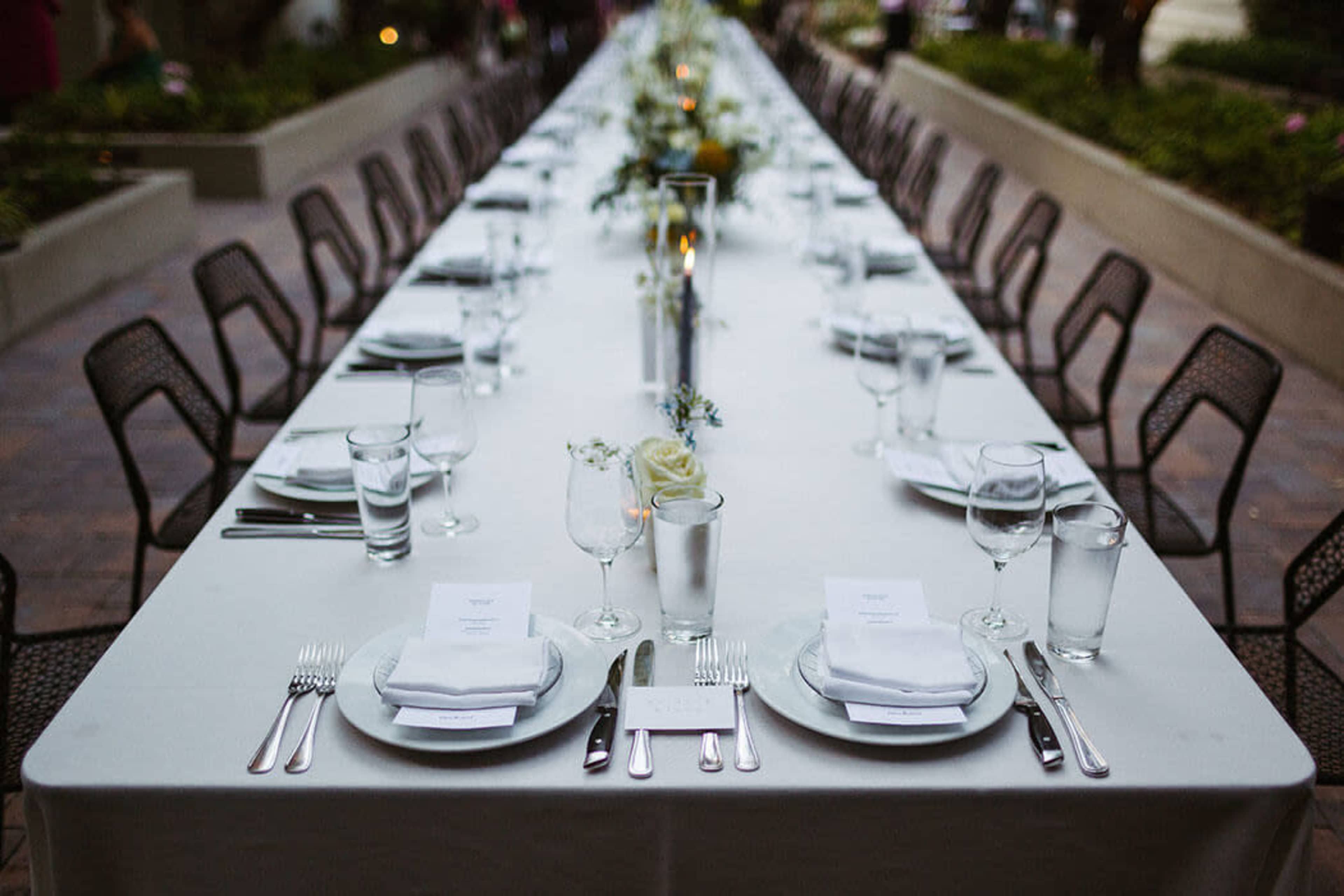 A long outdoor dining table is set with white plates, silverware, and glasses, adorned with a floral centerpiece and lit candles.