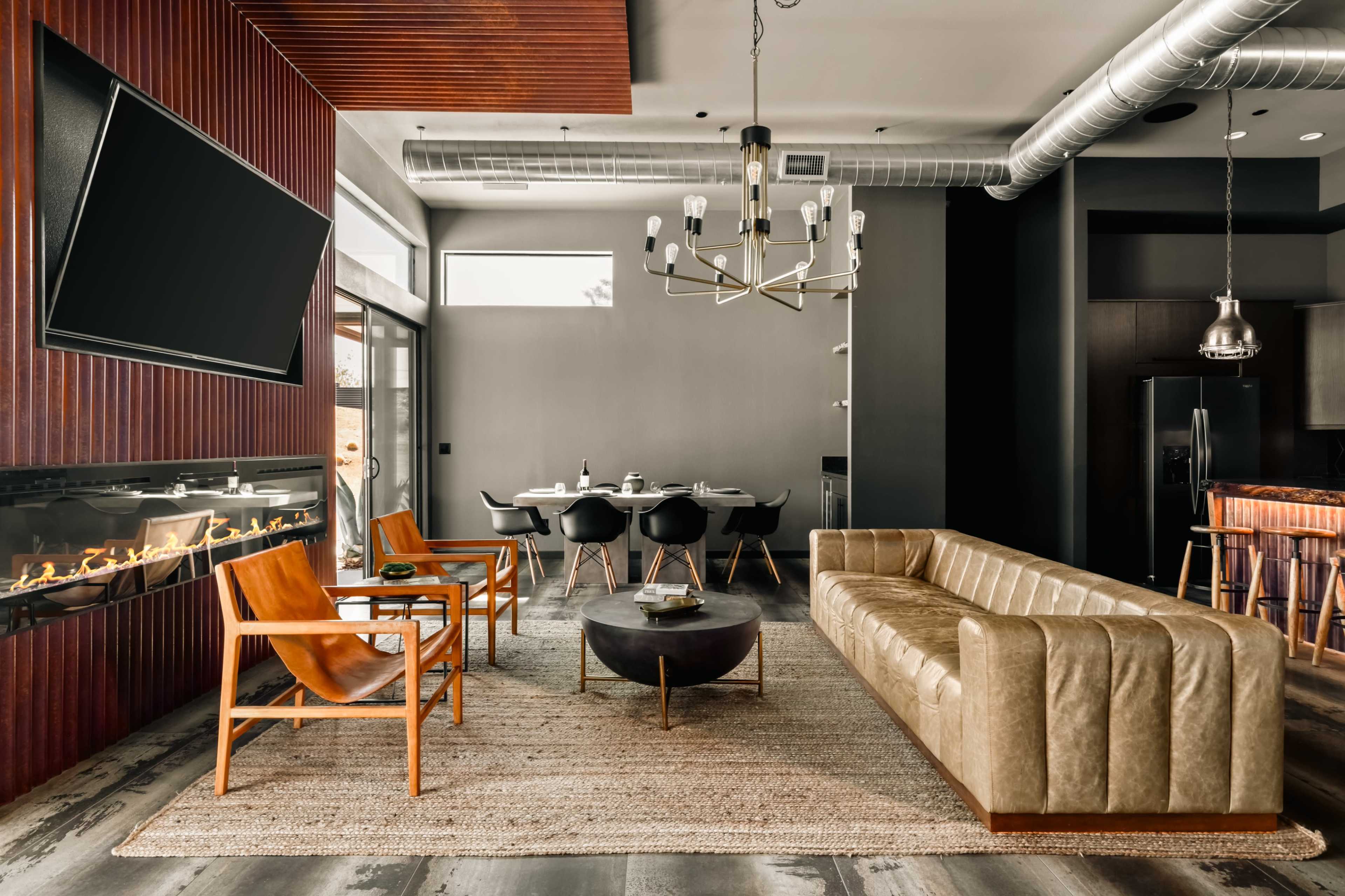 The image shows a modern living space featuring a leather sofa, two wooden chairs, a round coffee table, and a dining area with a circular table and chairs, all arranged around a sleek fireplace and large windows.