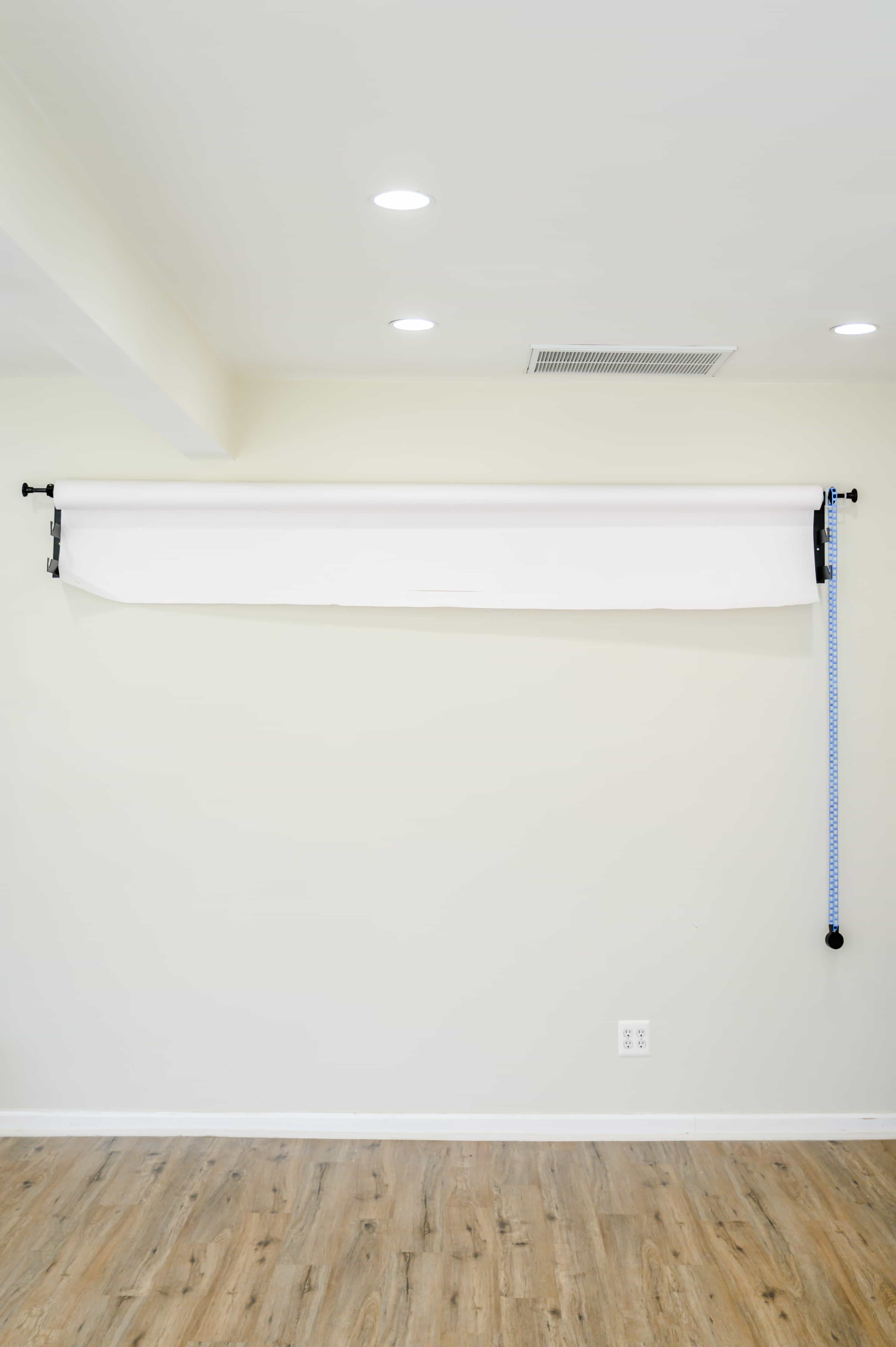 The image shows a blank wall with a roll-up white backdrop mounted near the ceiling, and a measuring tape hanging beside it.