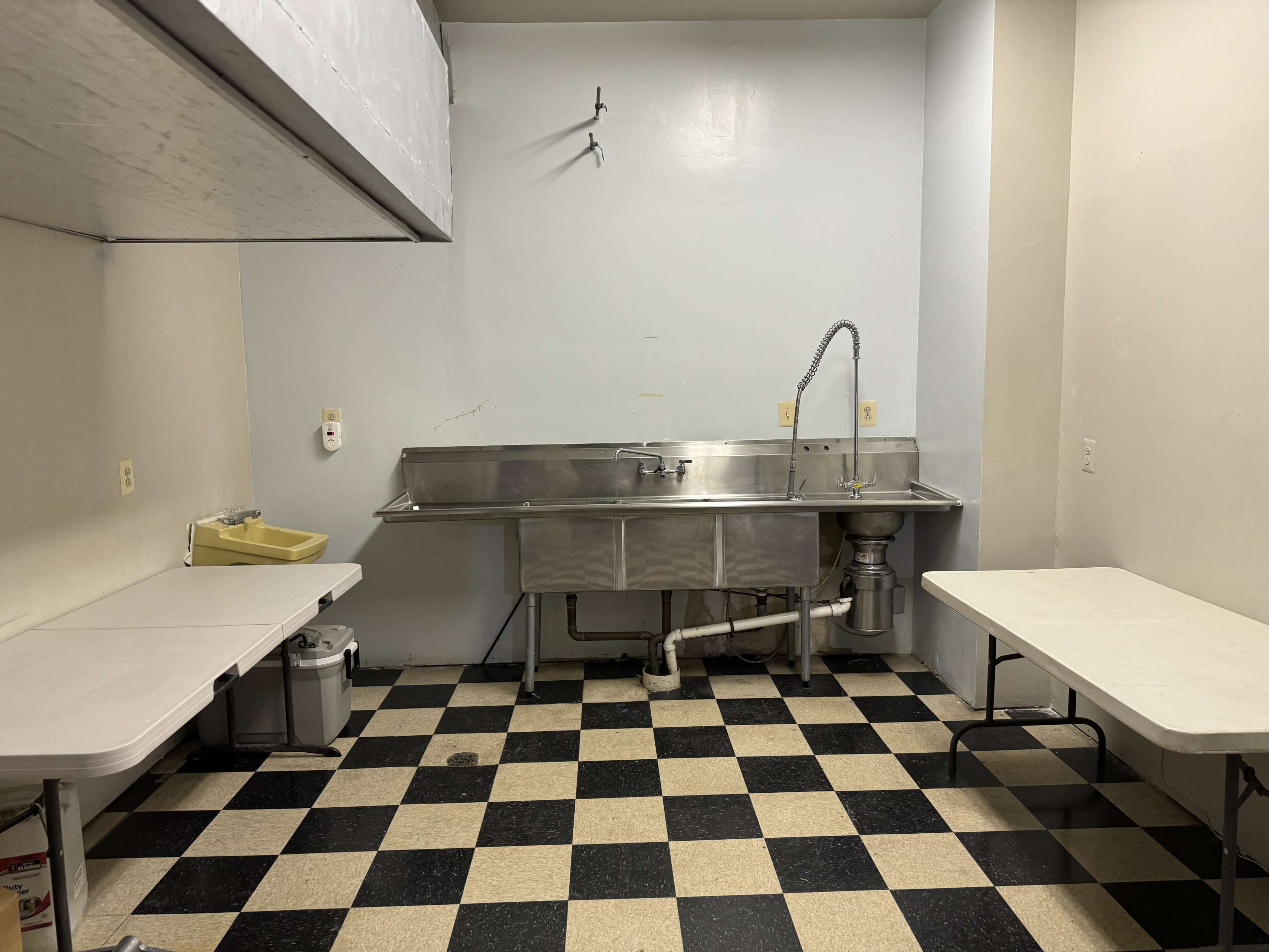 A small commercial kitchen space with a stainless steel sink, a couple of white tables, and checkered black and beige flooring.