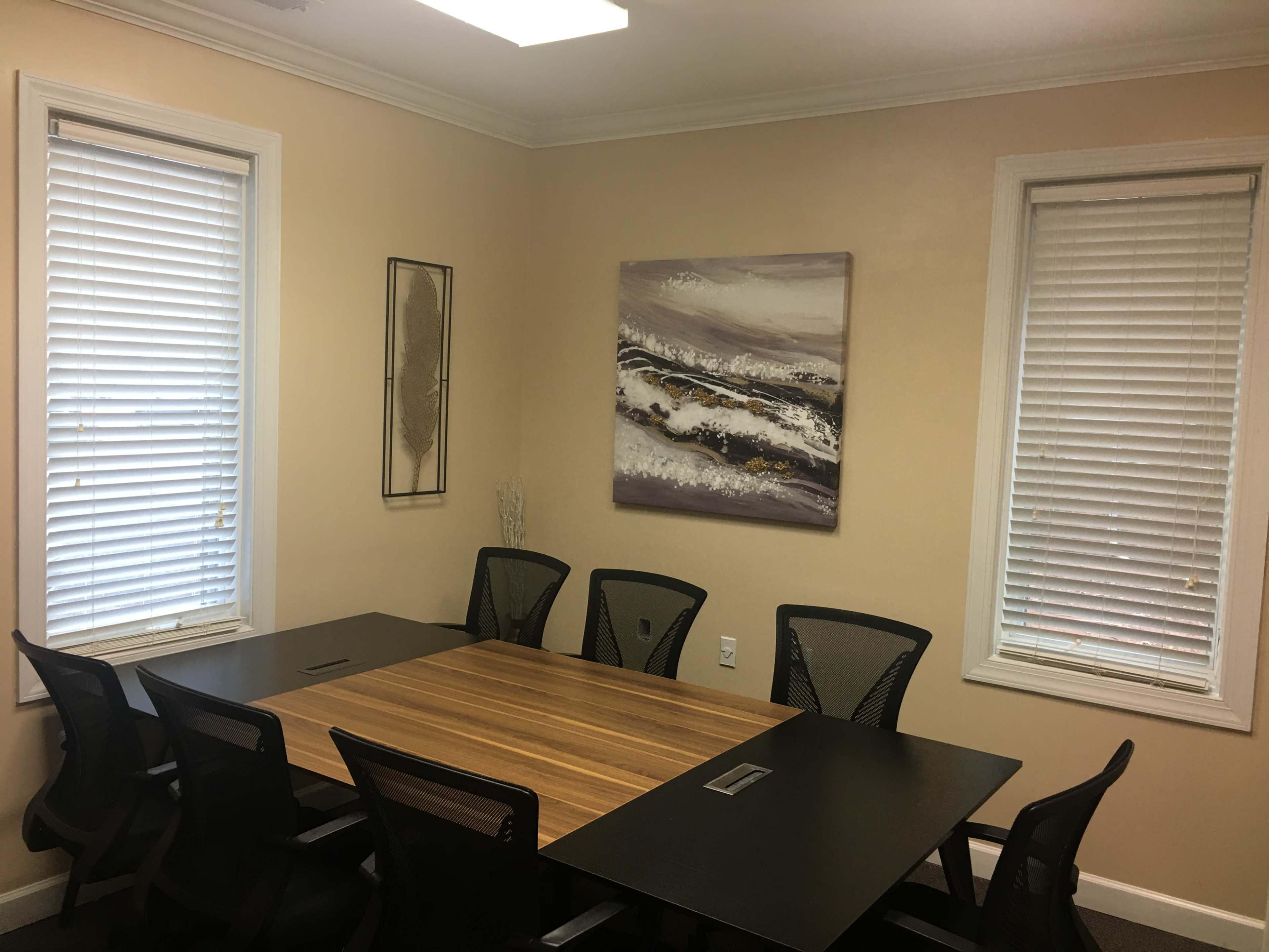 A small meeting room features a large wooden table surrounded by black mesh chairs, with a painting of abstract waves on the wall.