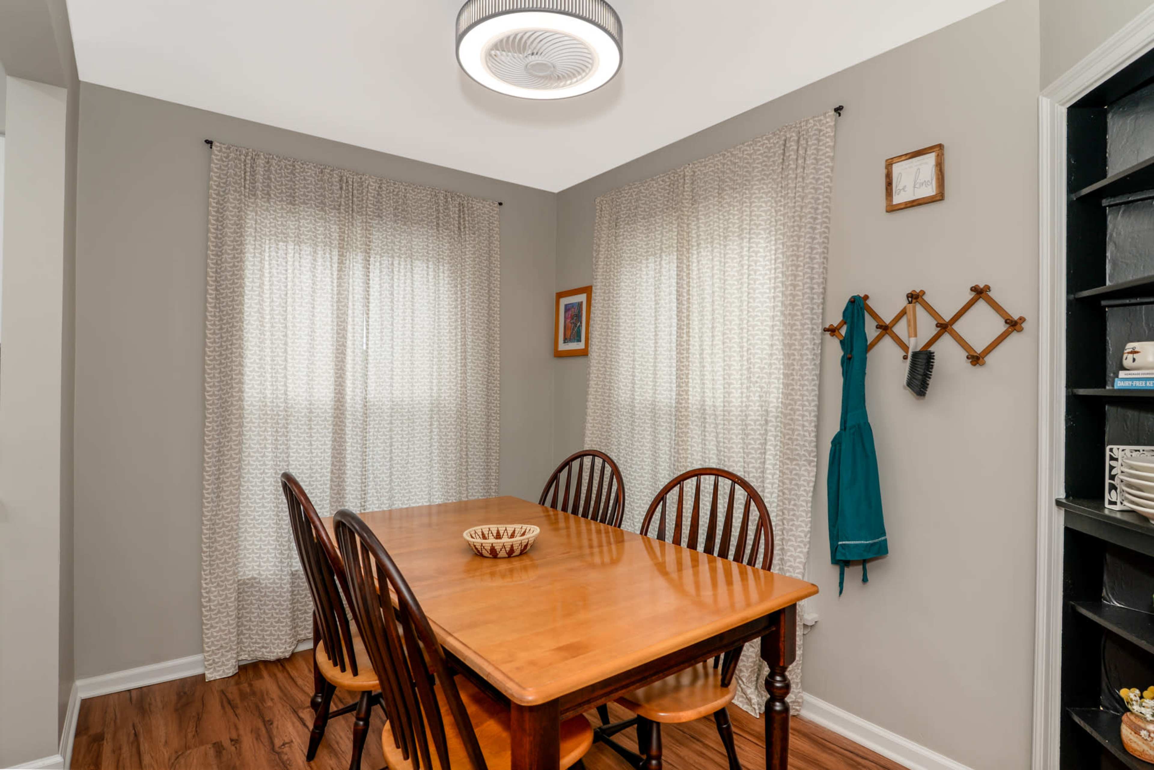 A wooden dining table surrounded by chairs is positioned in a well-lit room with sheer curtains and a shelf displaying various items.
