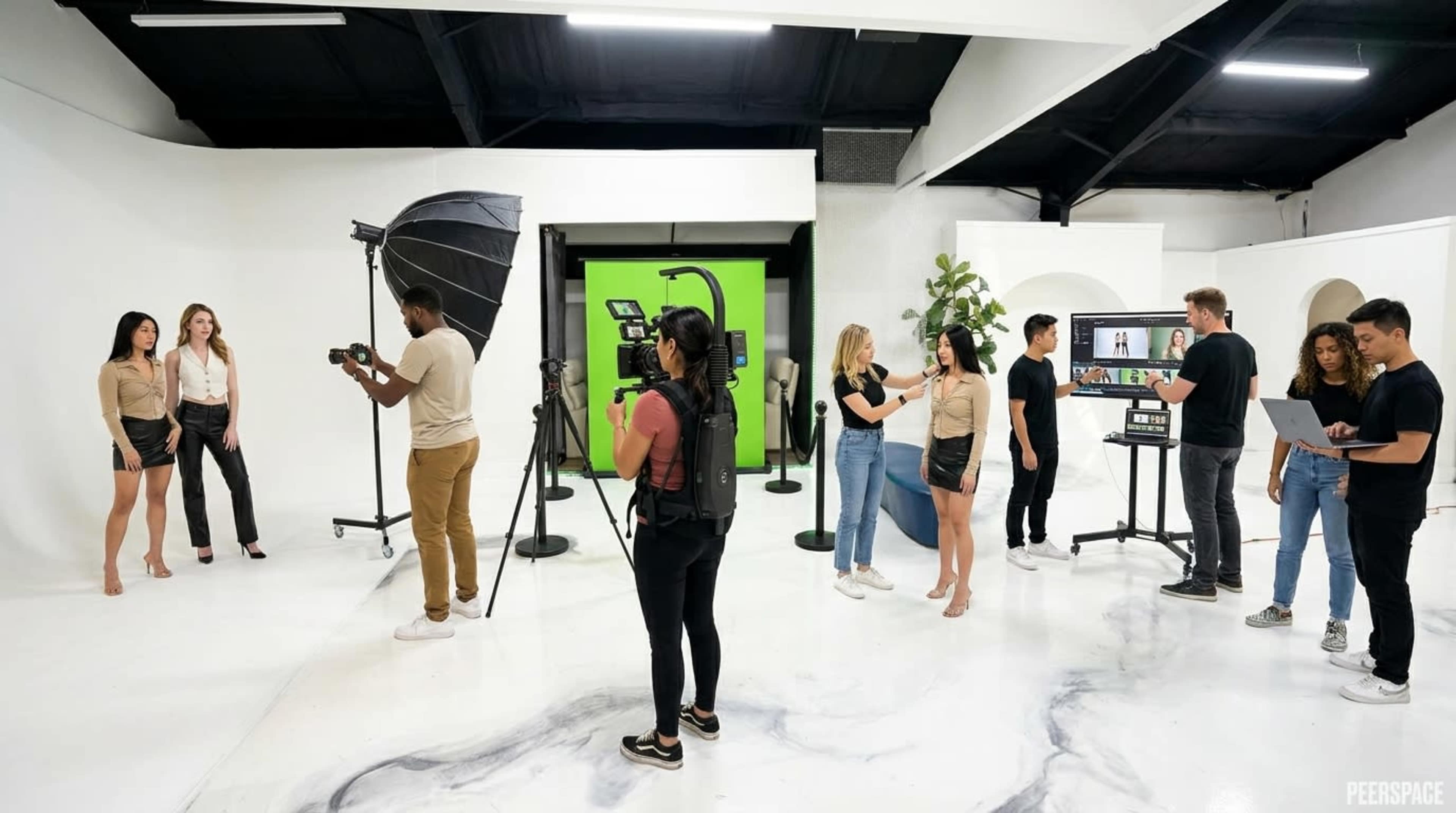 A group of people is engaged in a photoshoot in a studio with a white backdrop and a green screen, while others are operating cameras and monitors.