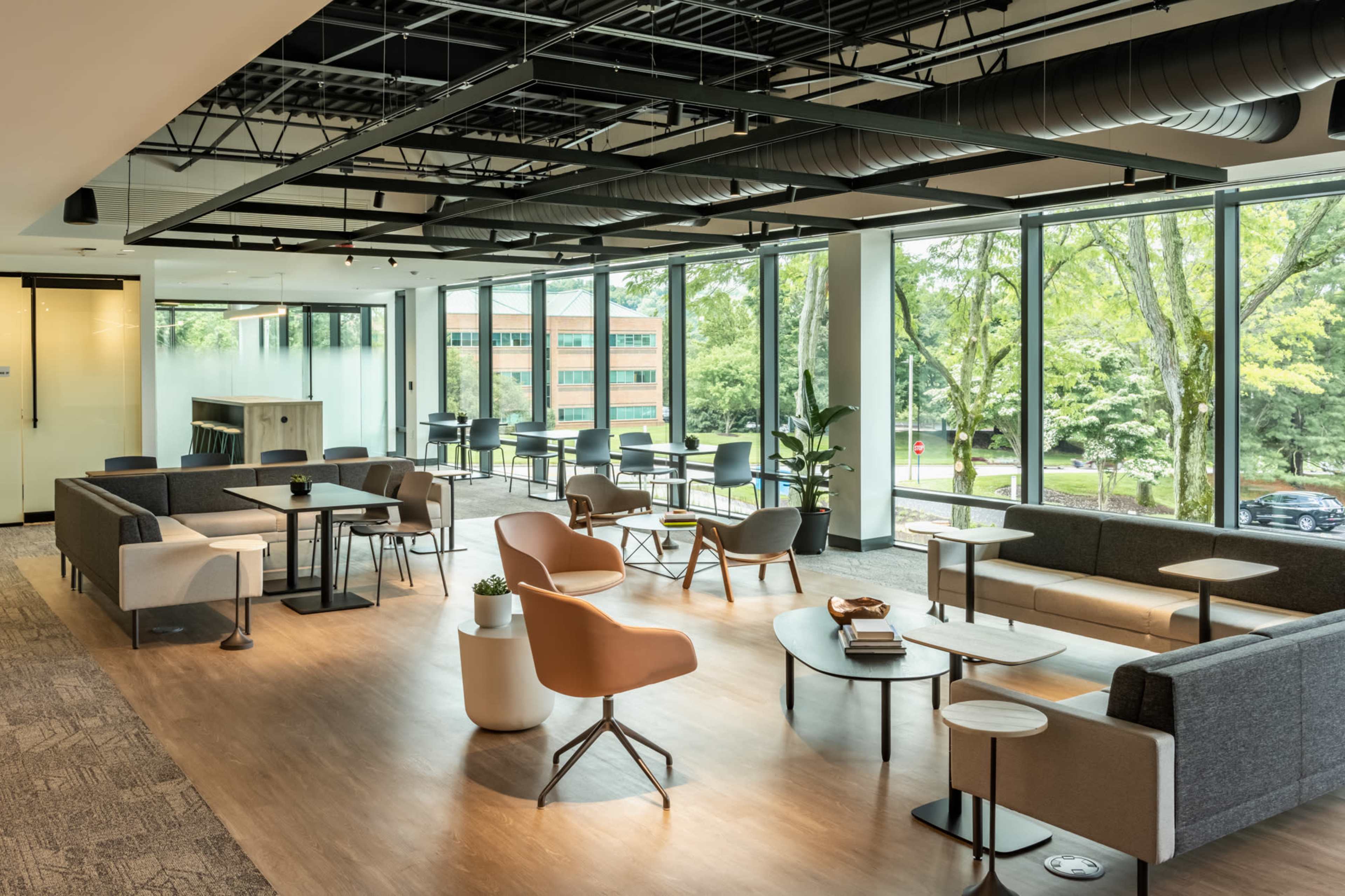 A modern office lounge area with various seating arrangements, large windows allowing natural light, and a view of greenery outside.