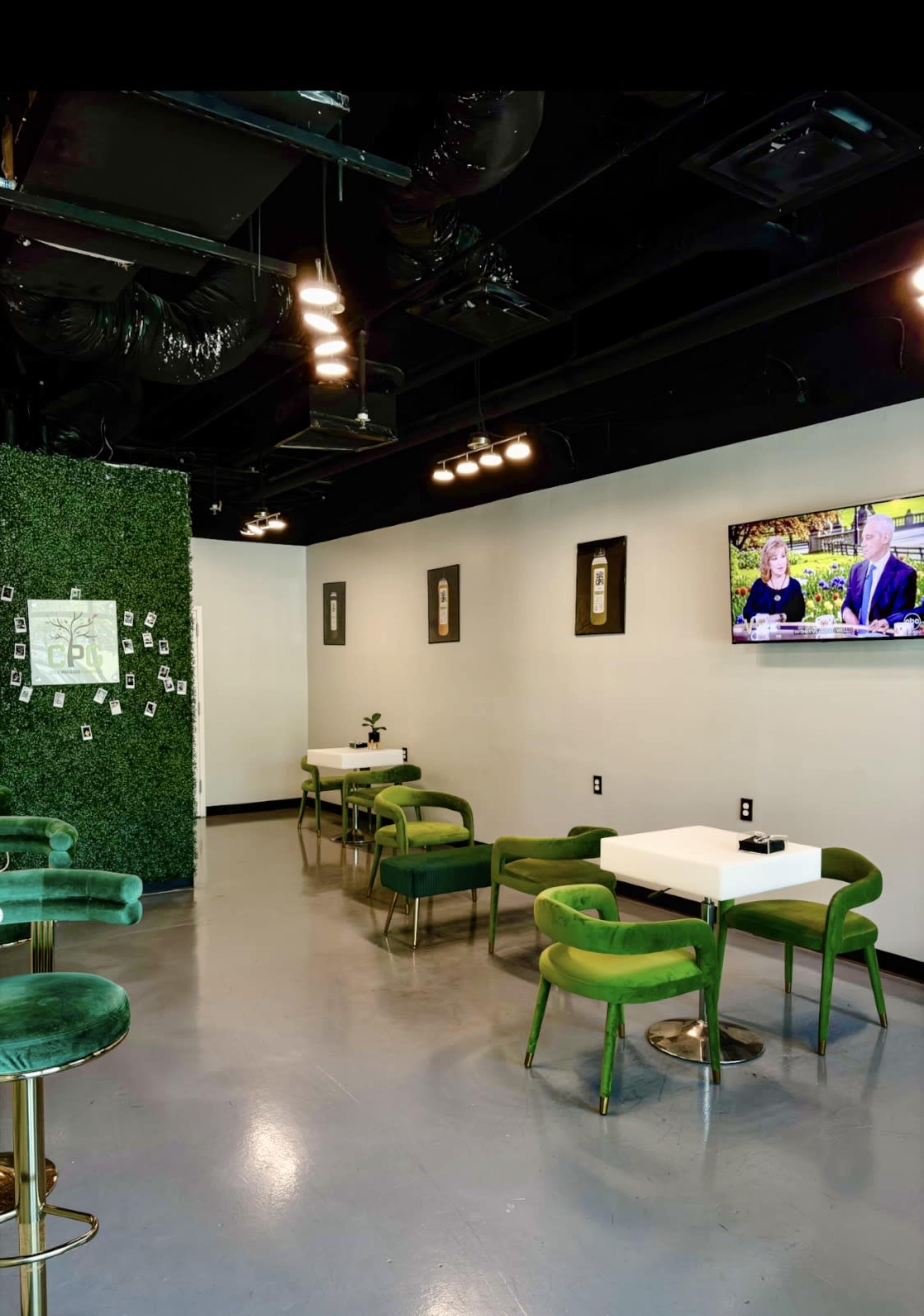 The image shows a modern cafe interior featuring green velvet chairs and tables, illuminated by ceiling lights, with a television mounted on the wall and decorative artwork.