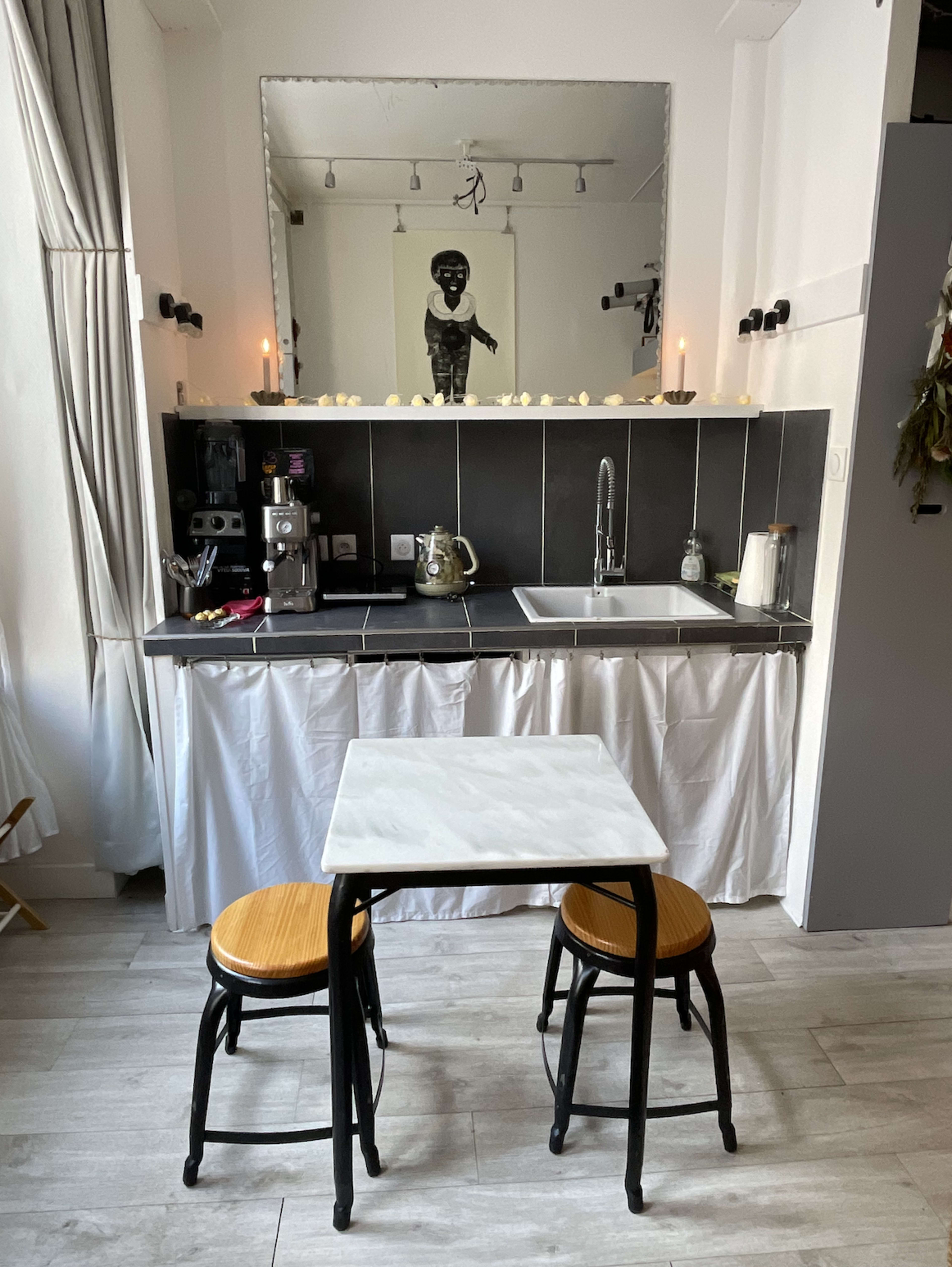 A small kitchen features a black countertop with a sink, coffee machine, and a table set for two with bamboo chairs beneath a large mirror.