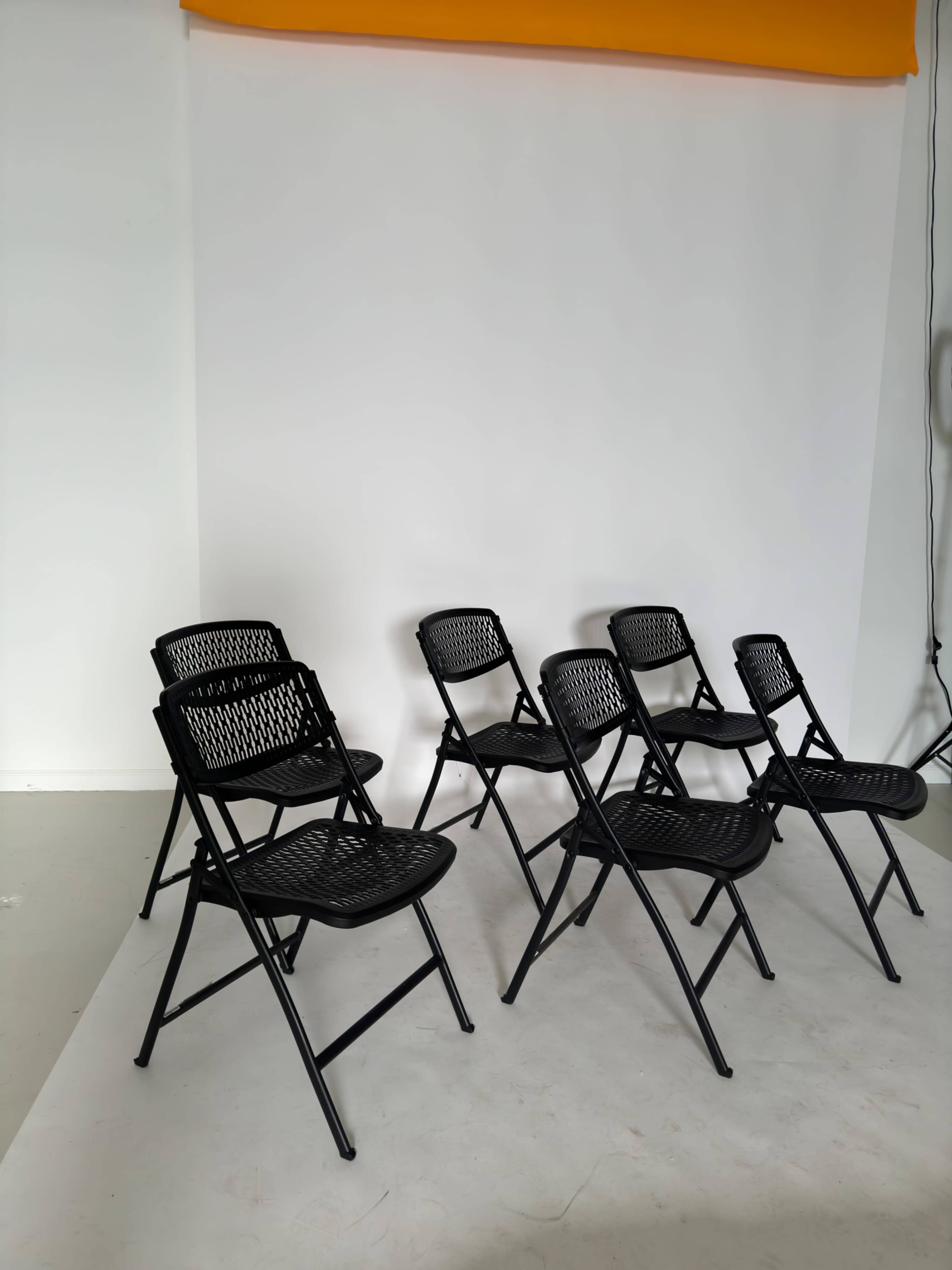 A set of six black folding chairs is arranged on a light-colored floor in front of a plain white backdrop.