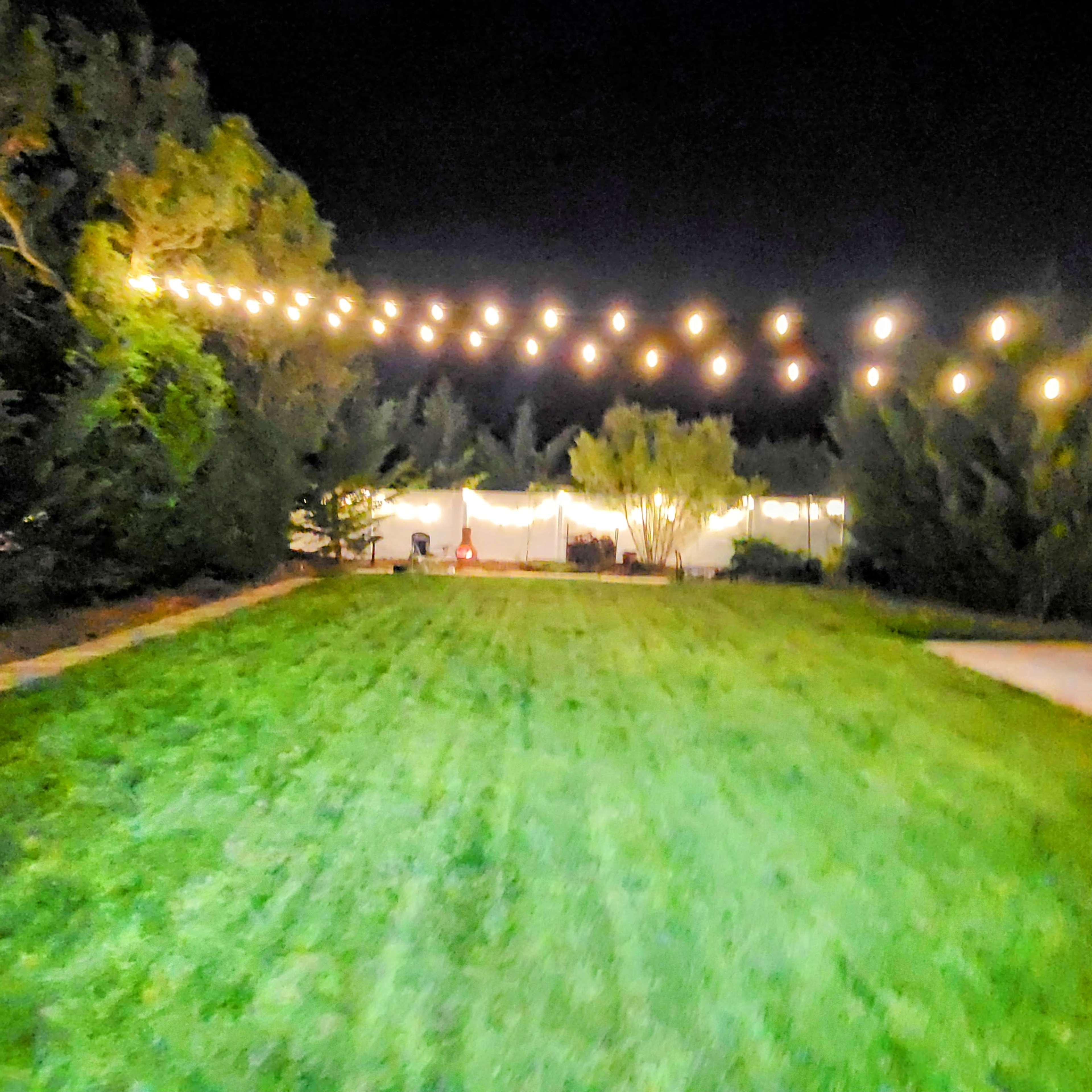 A well-lit outdoor area features string lights overhead, leading to a tent or gathering space surrounded by trees and a grassy lawn at night.