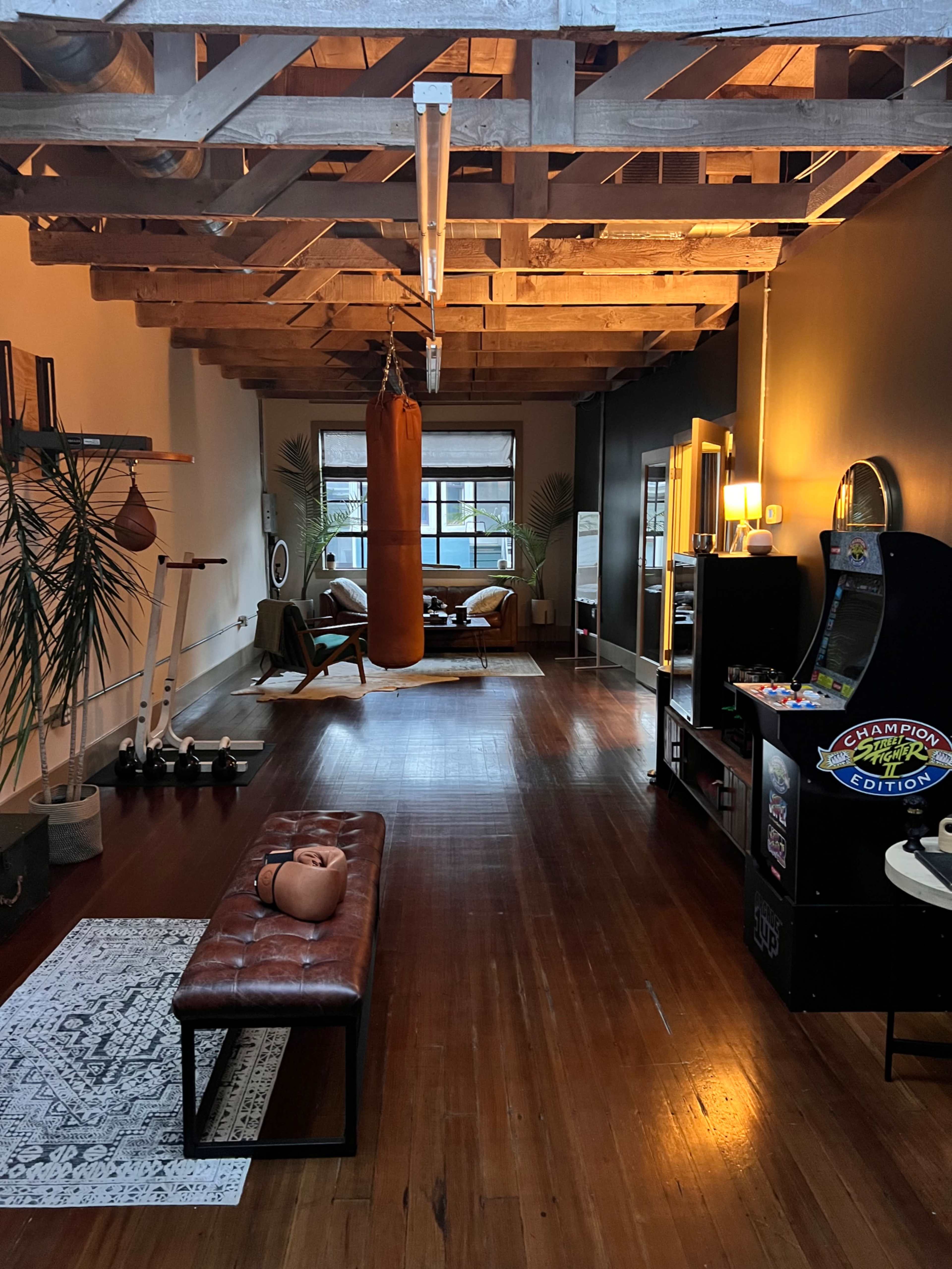 An open, spacious room with wooden beams, featuring a punching bag, a vintage arcade machine, a bench, and a lounge area with a couch and plants.