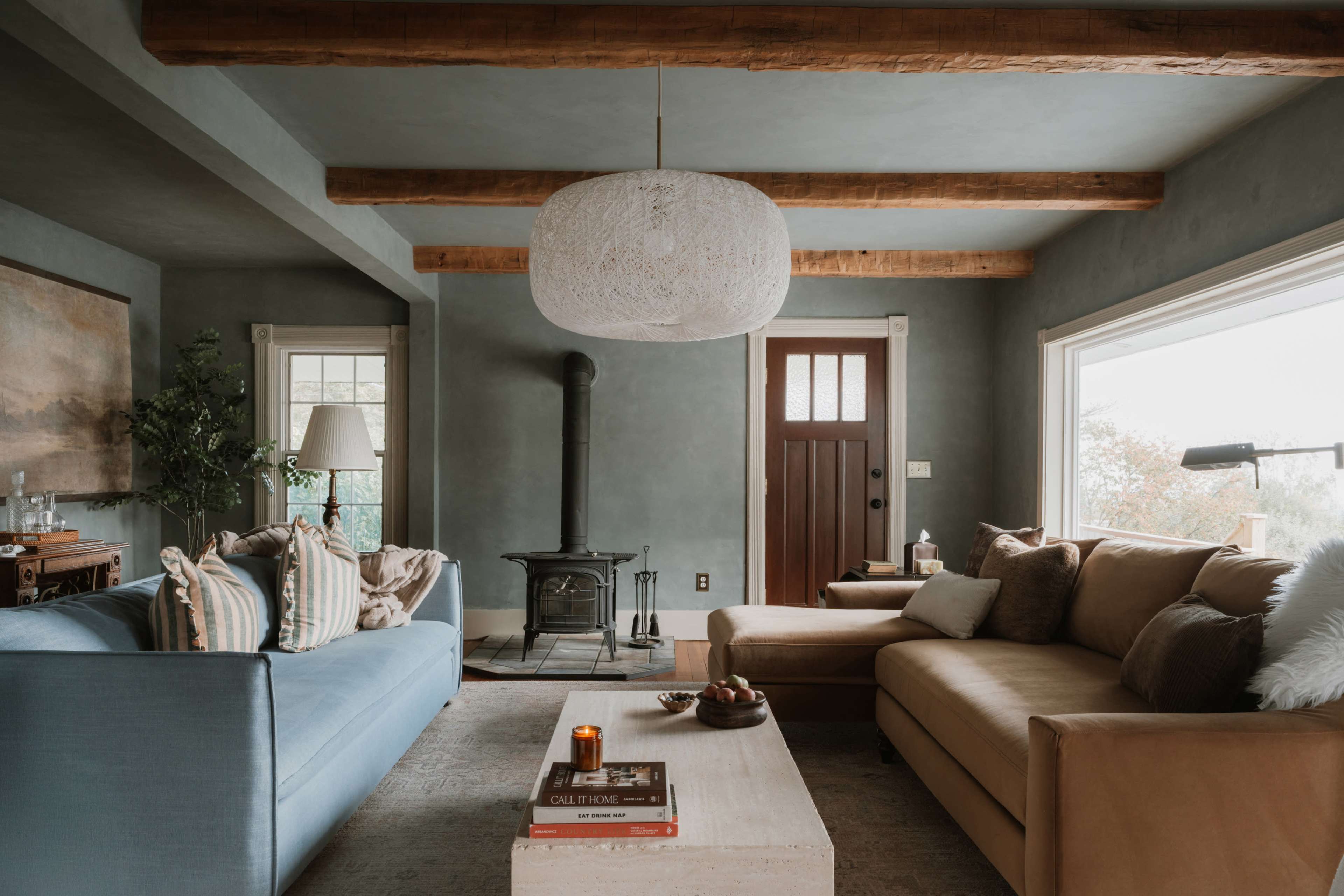 The cozy living room features a blue sofa, a tan sectional couch, a wooden coffee table, and a wood-burning stove, with large windows allowing natural light.