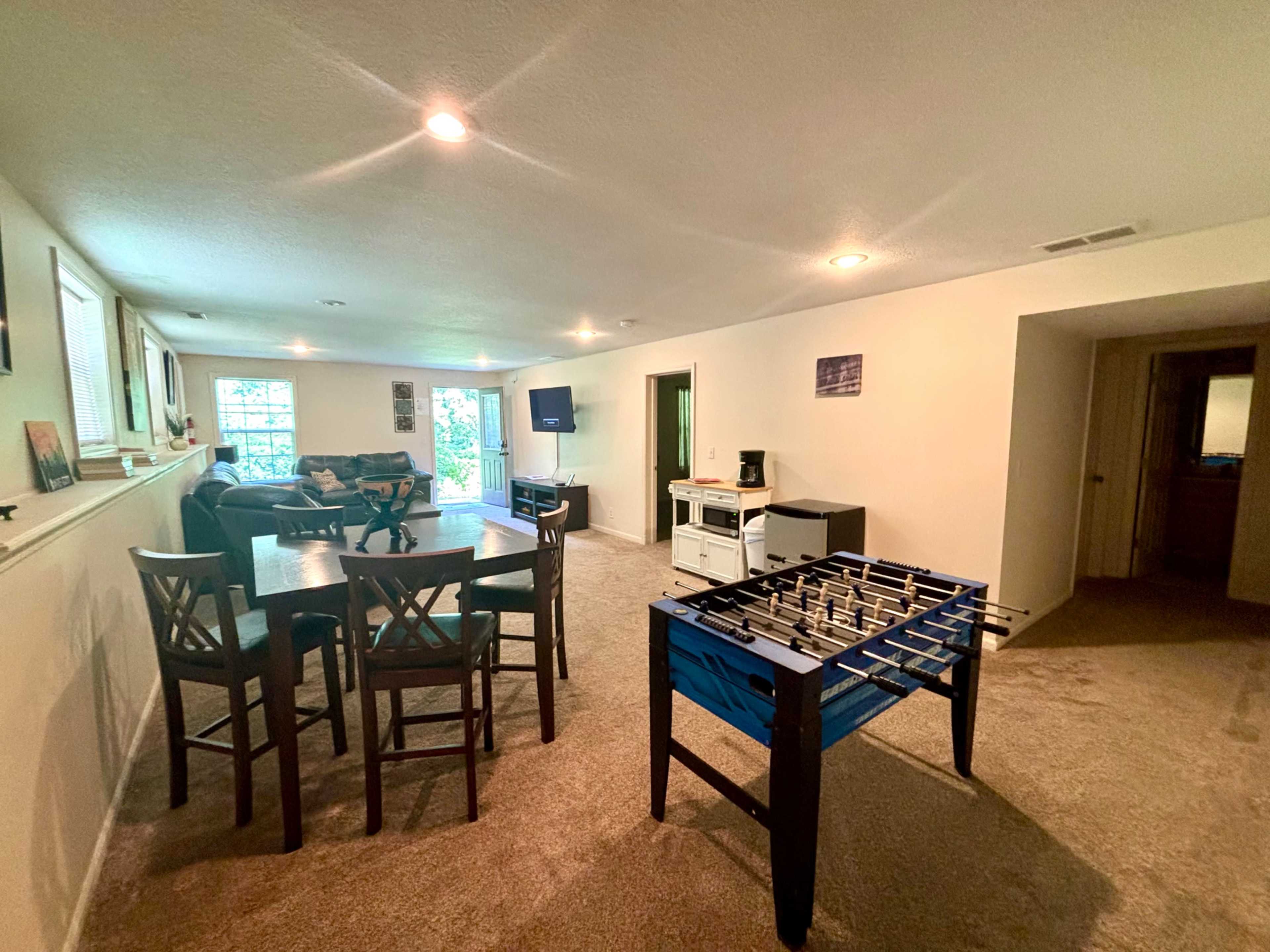 The image shows a spacious basement room featuring a foosball table, seating area, and a small kitchenette.