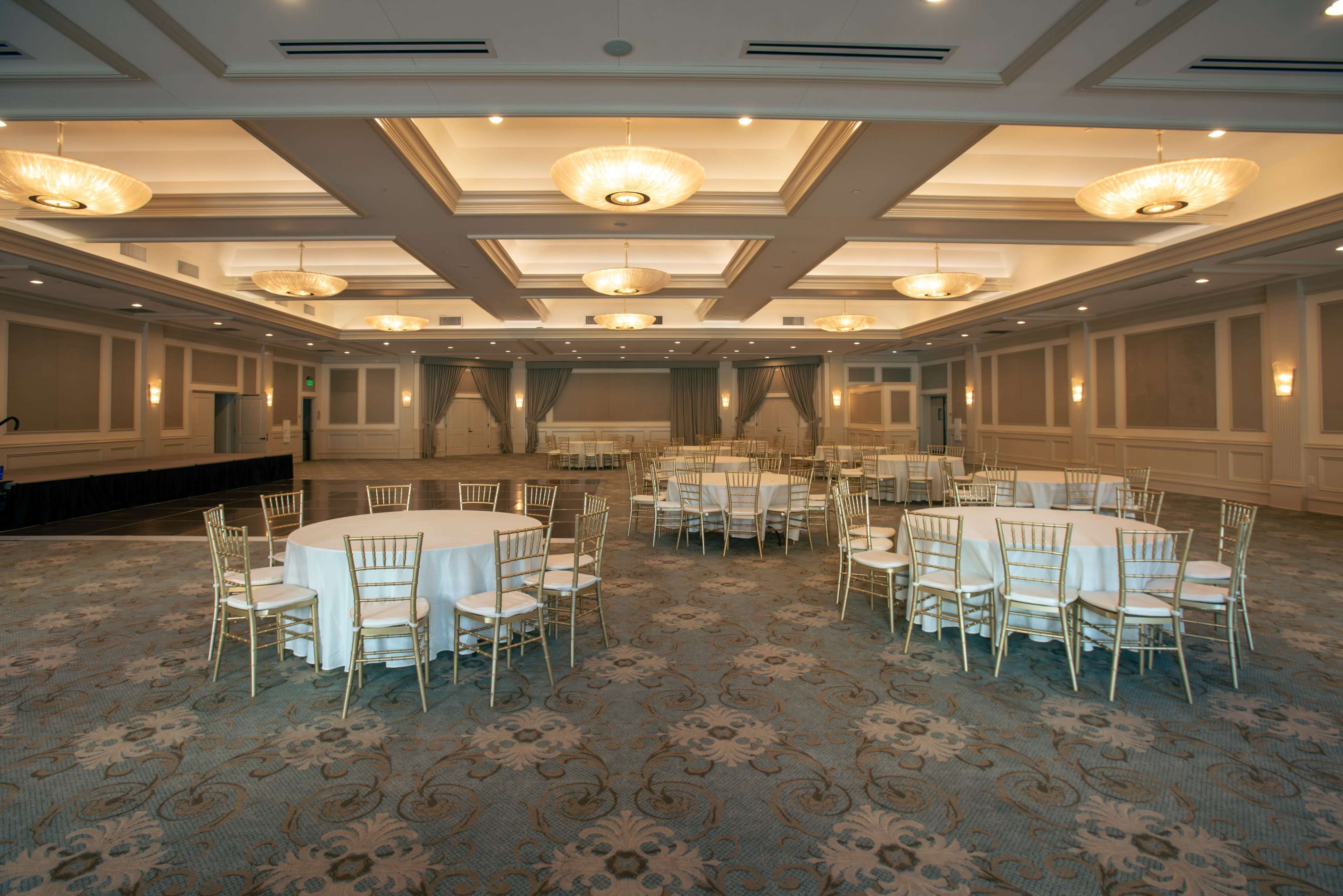 The image shows a spacious event room with round tables covered in white tablecloths and gold chairs arranged throughout the area.