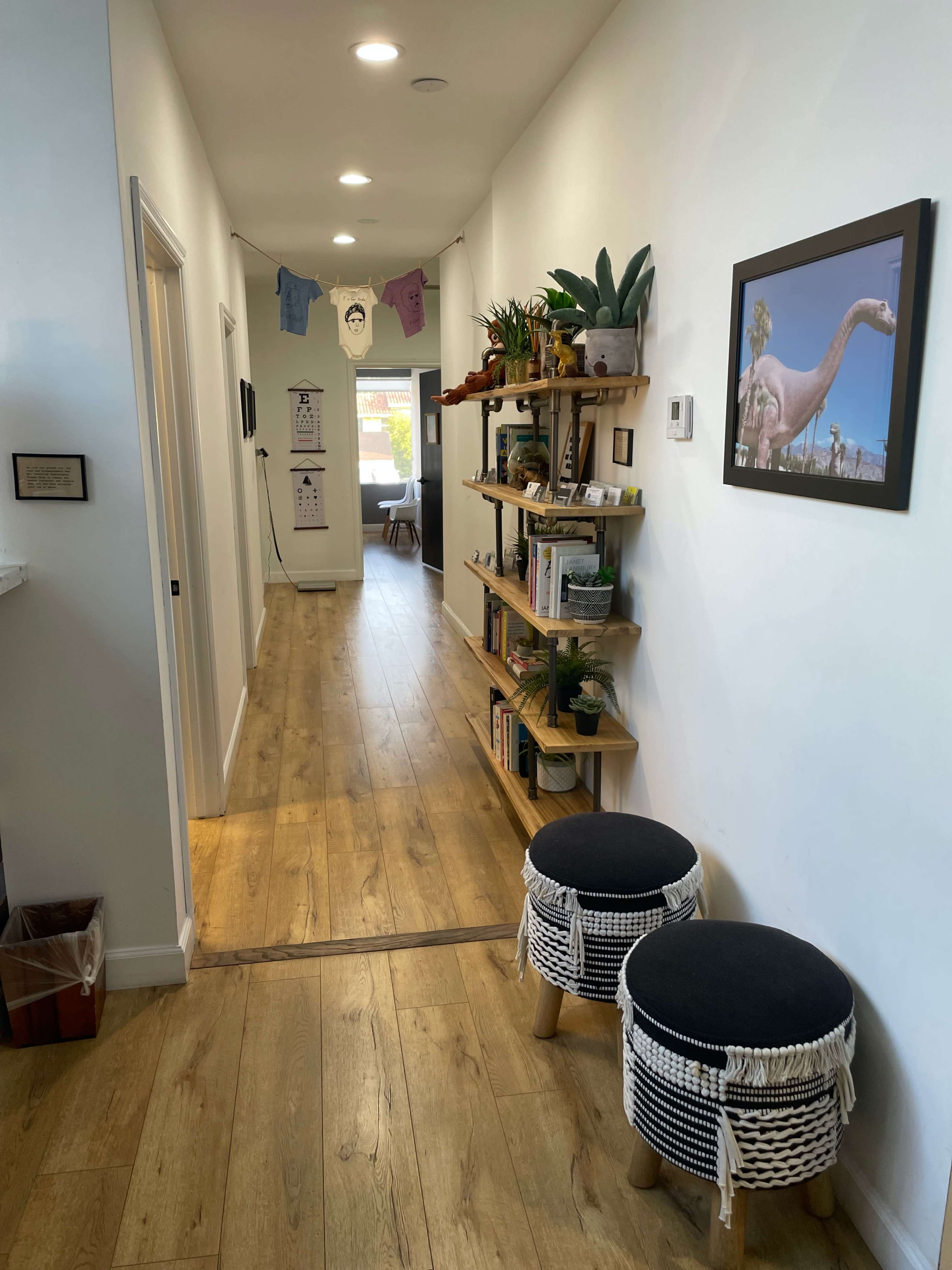 A bright hallway with light wood flooring, a shelf filled with plants and books on one side, and two small stools on the floor opposite a wall adorned with a framed picture.