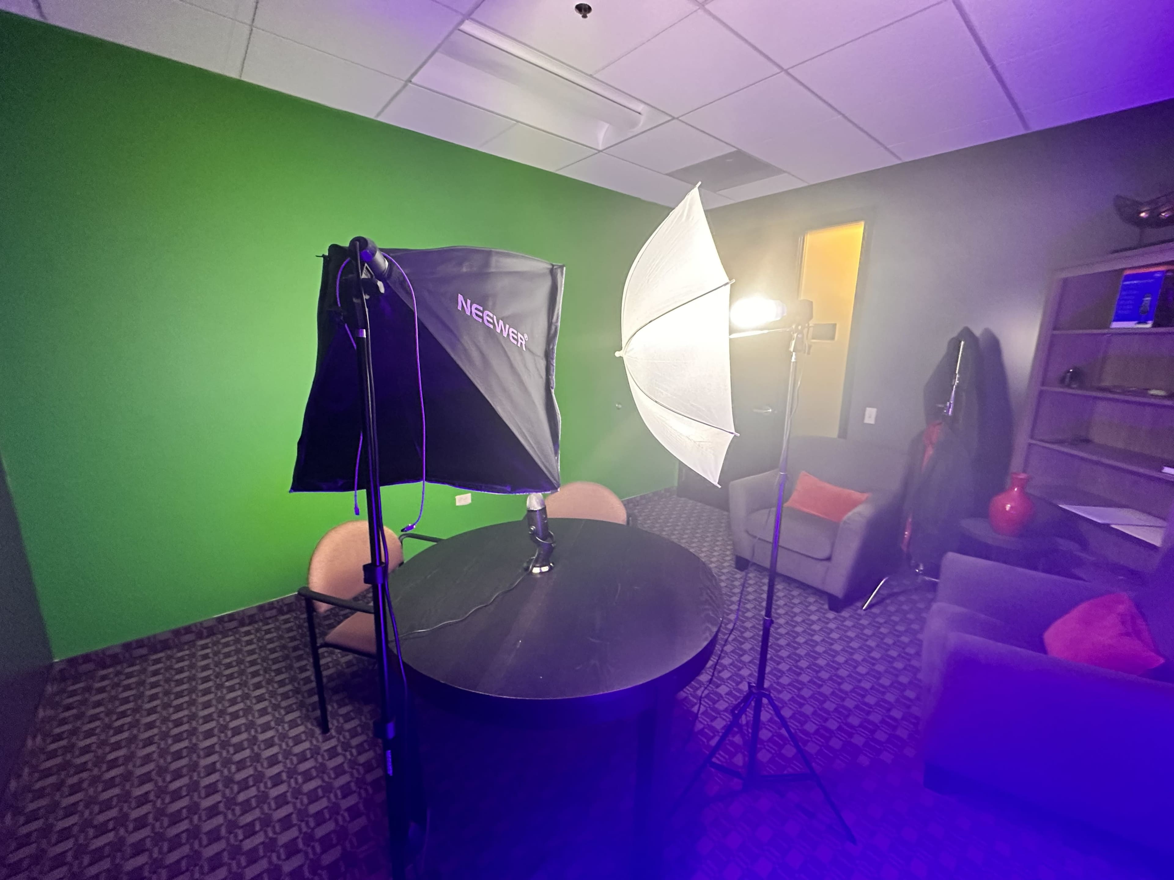 A small room with a round table, two chairs, and lighting equipment set up against a green screen backdrop.
