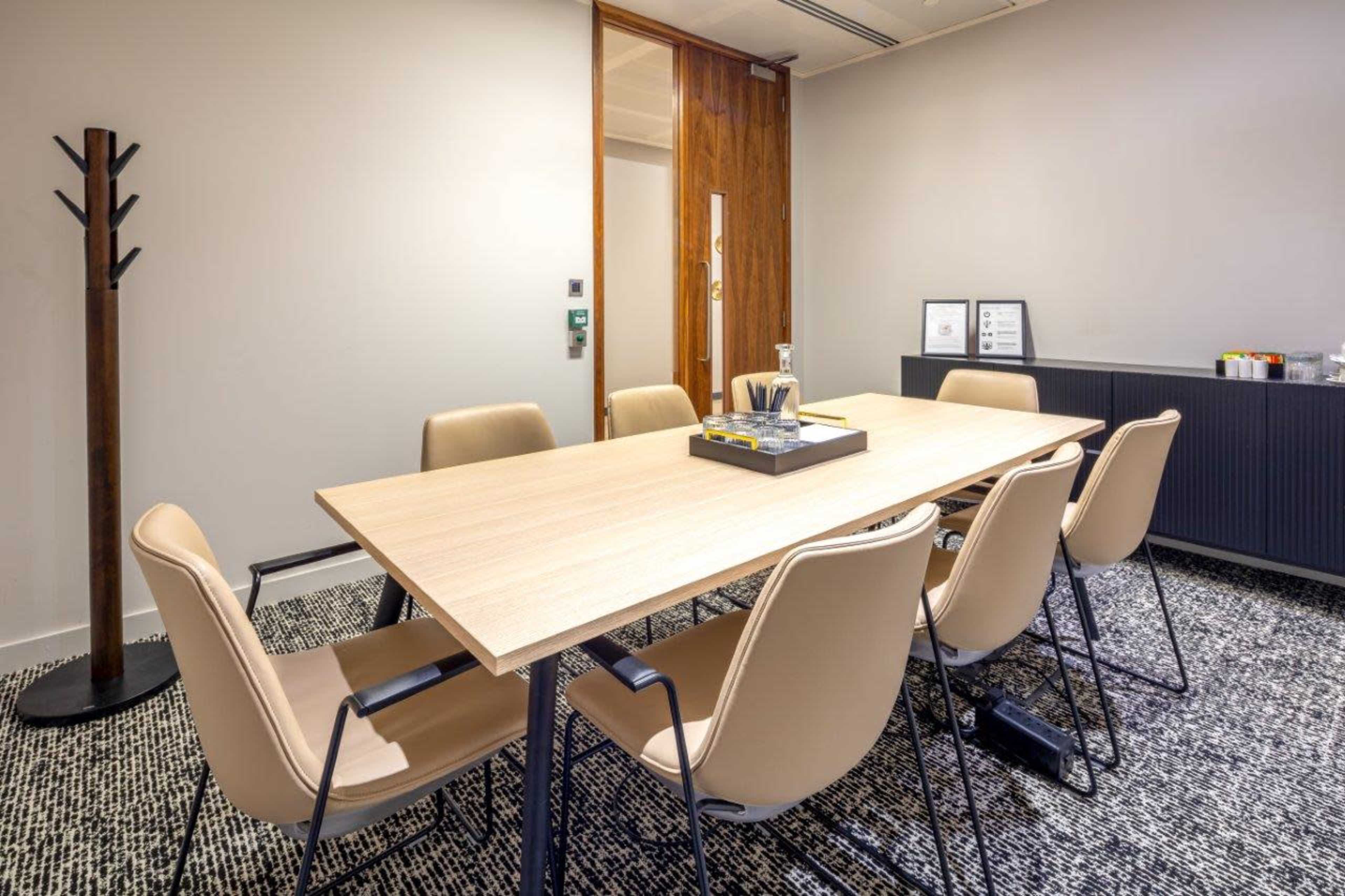 A modern meeting room features a long wooden table surrounded by eight beige chairs, with a coat rack and a textured rug on the floor.