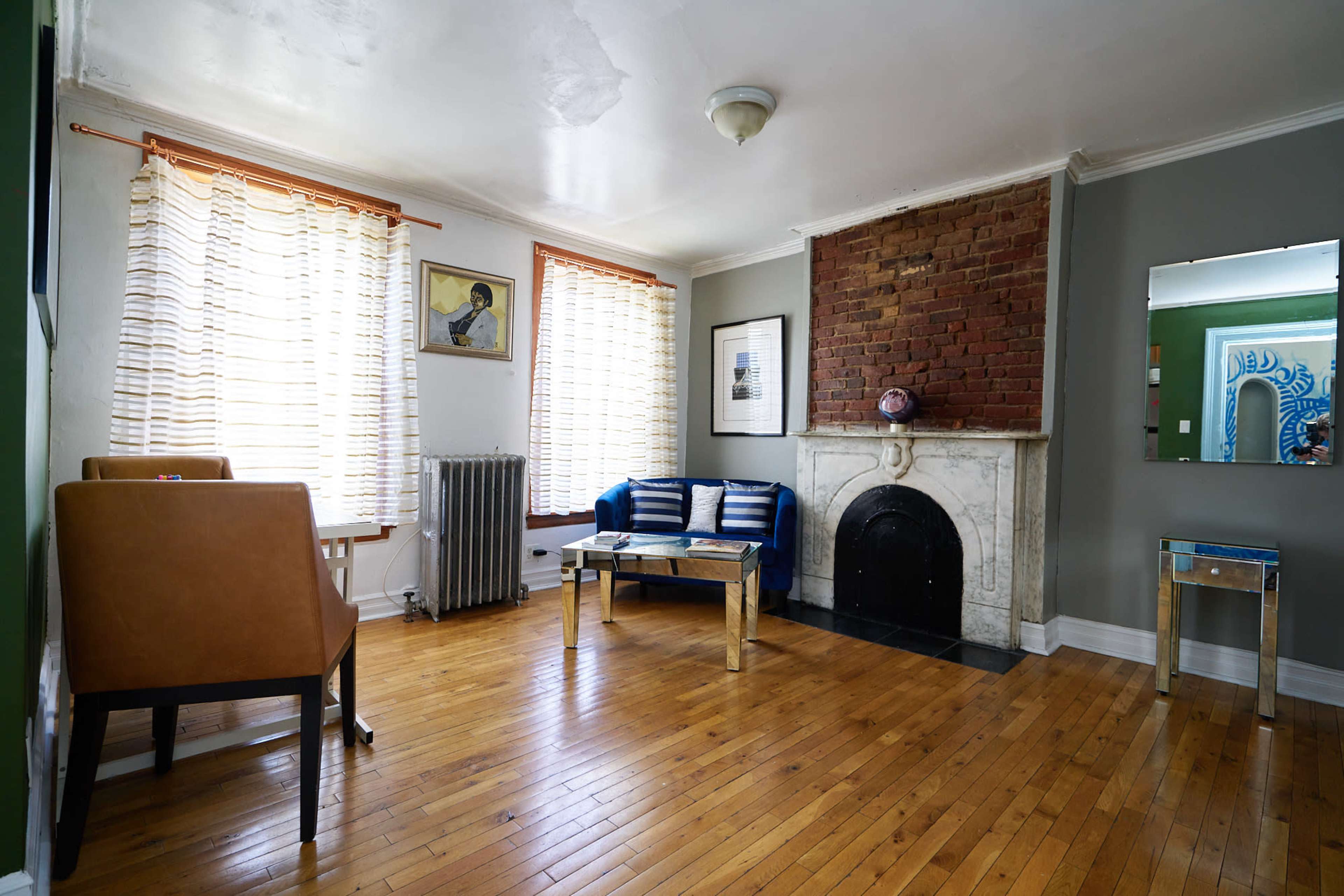 The image shows a living room with hardwood floors, a brick fireplace, two chairs, and large windows with sheer curtains.