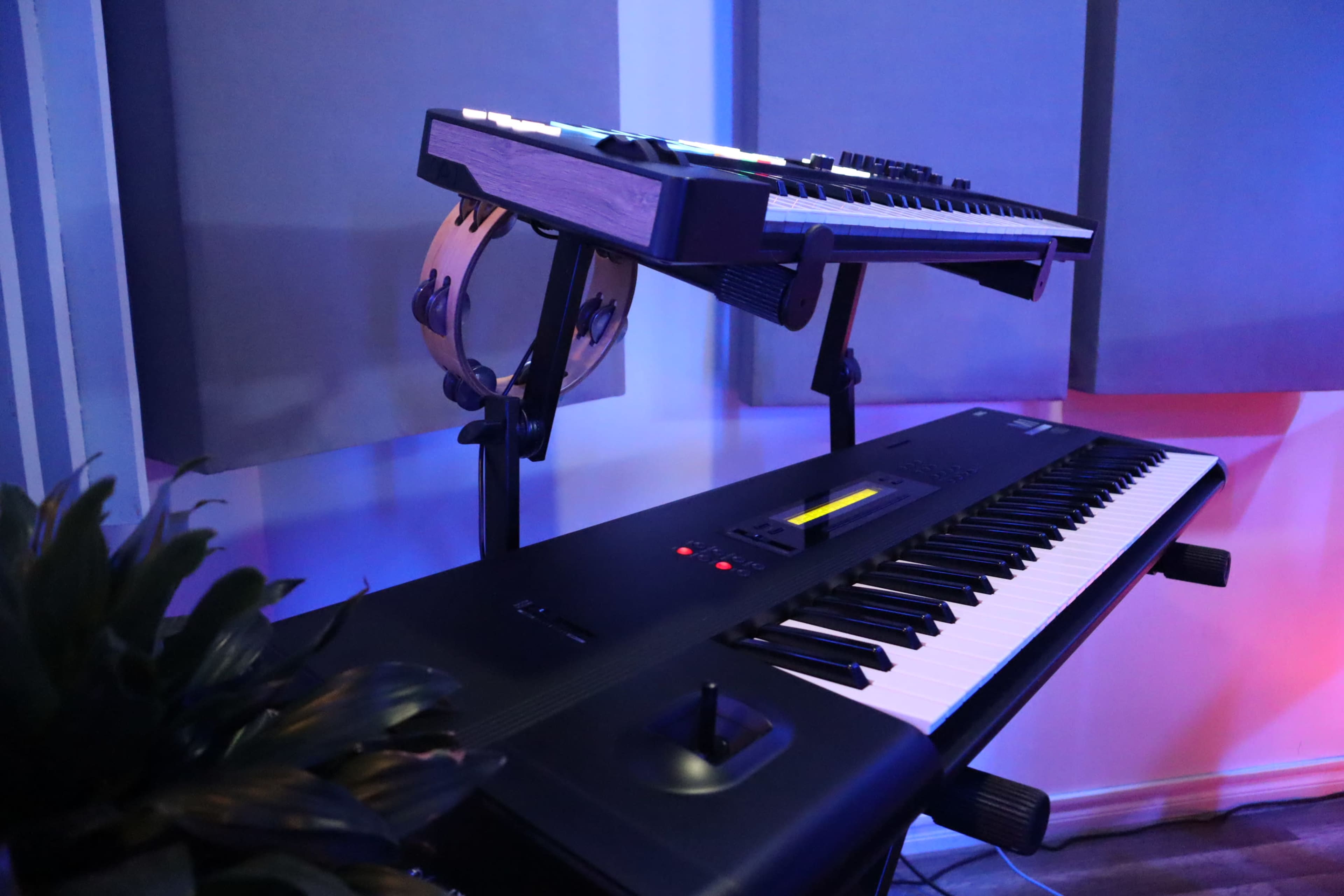 A digital piano is positioned below a synthesizer on a stand, with a tambourine nearby in a studio setting.