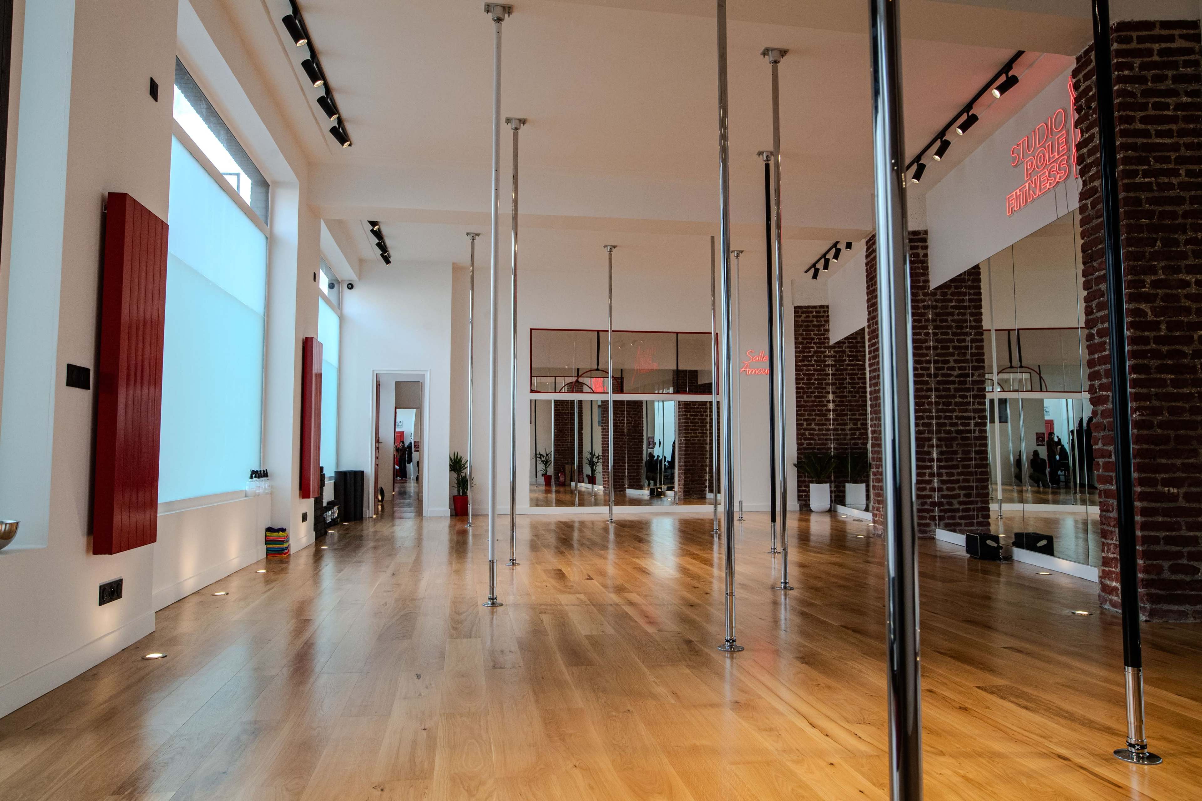 The image shows a spacious studio with wooden flooring, pole dance poles arranged evenly, and large windows allowing natural light to enter.