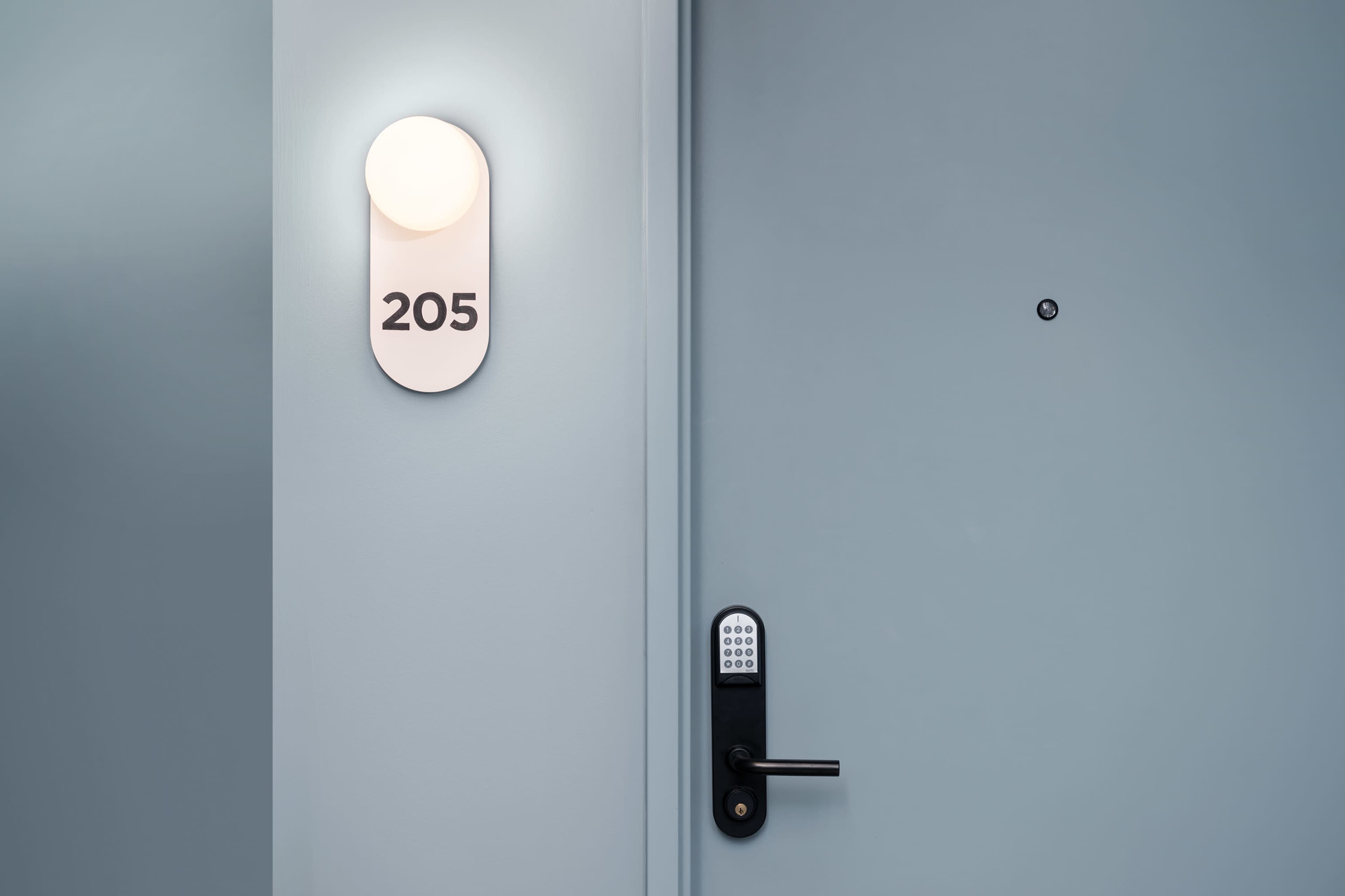A hotel room door labeled "205" is illuminated by a circular light on a pale blue wall.