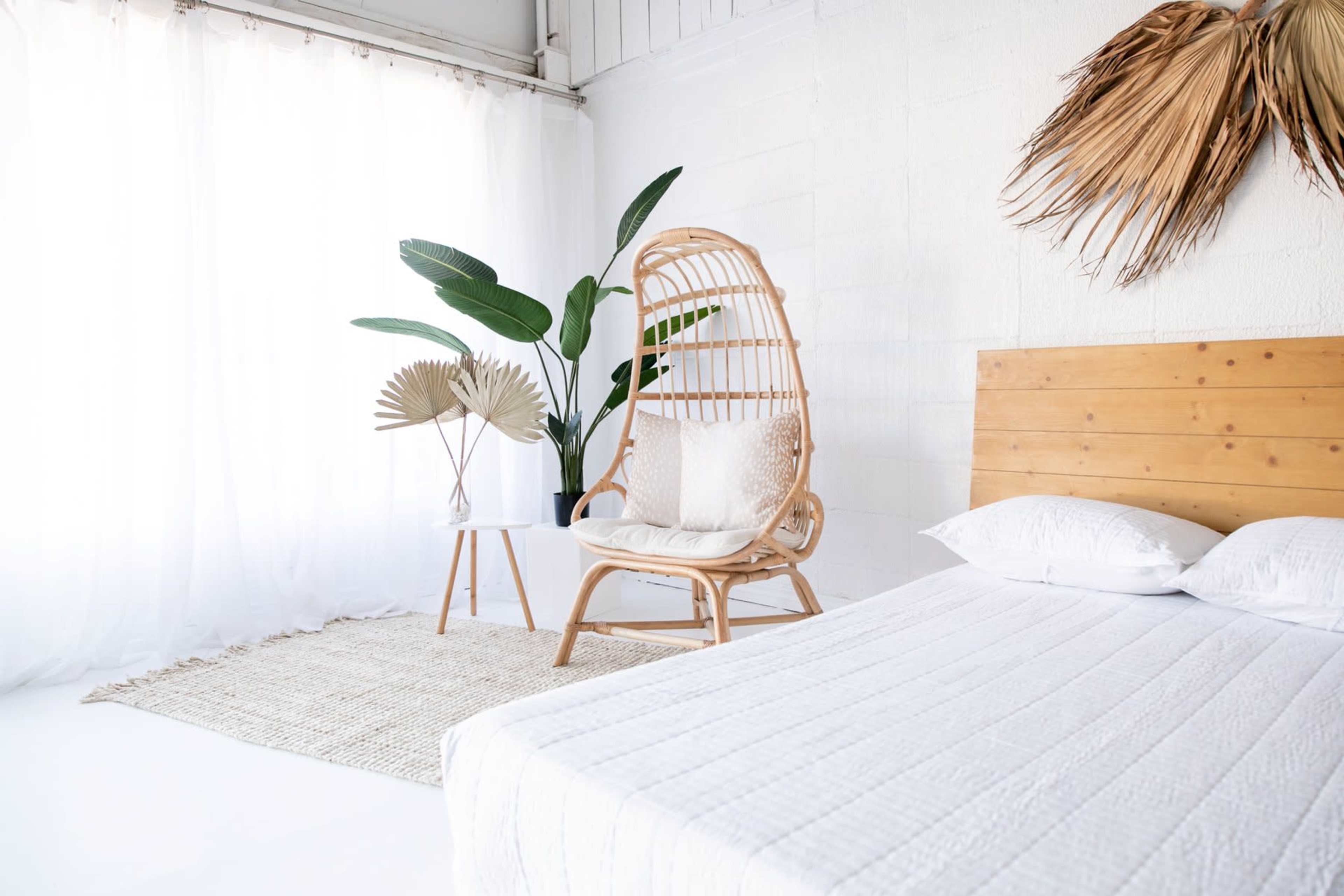A minimalist bedroom features a bed with a white quilt, a rattan chair, and potted plants beside a small table, all set against a backdrop of white walls and curtains.