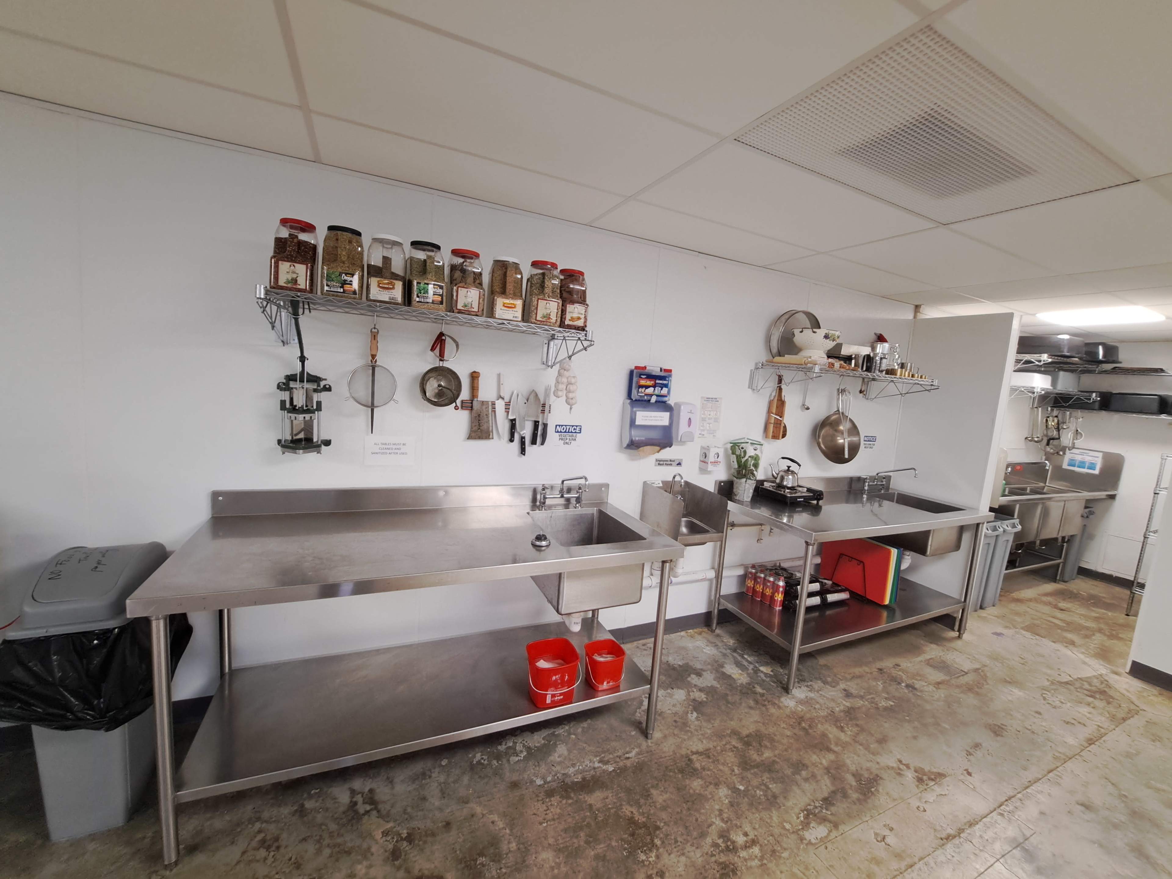 The image shows a clean commercial kitchen with stainless steel work tables, storage shelves, and various utensils and ingredients organized on the walls.