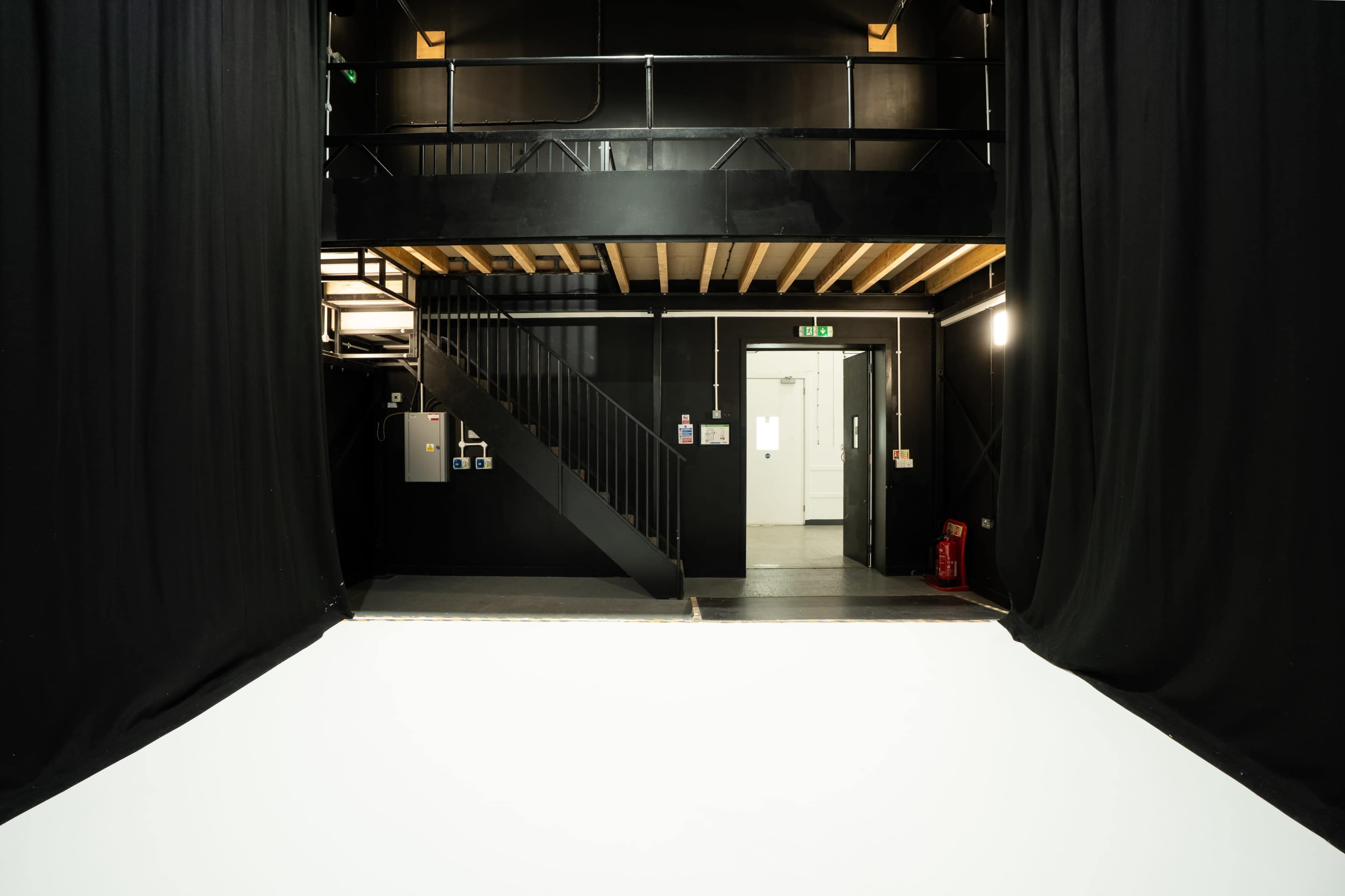 The image shows a large, empty studio space with black curtains, a staircase on the left, and a white floor area leading to a door at the back.
