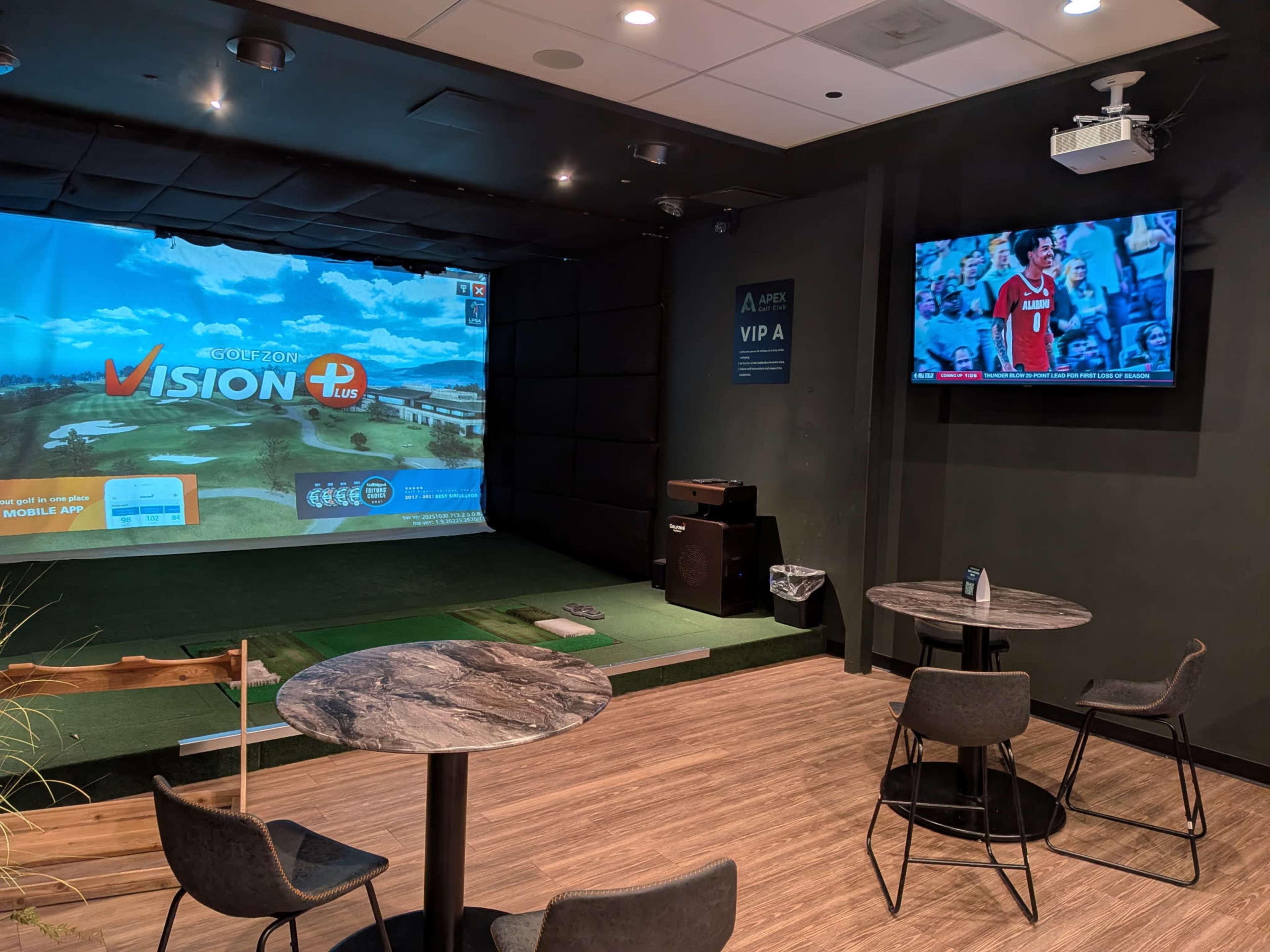 The image shows an indoor golf simulator area with a large screen displaying a virtual course and a television showing a sports event, accompanied by small tables and chairs.
