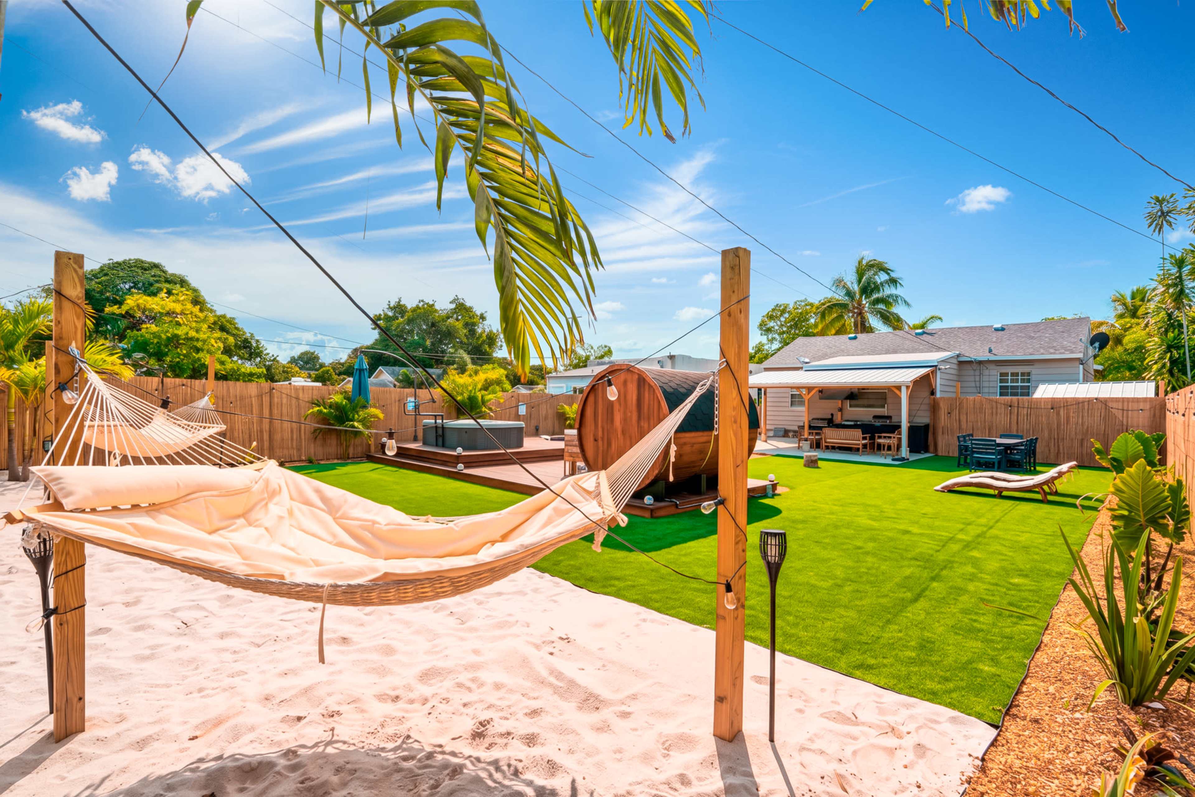 The image shows a backyard with a sandy area featuring a hammock, artificial grass, a wooden barrel sauna, and a seating area surrounded by palm trees and a wooden fence.